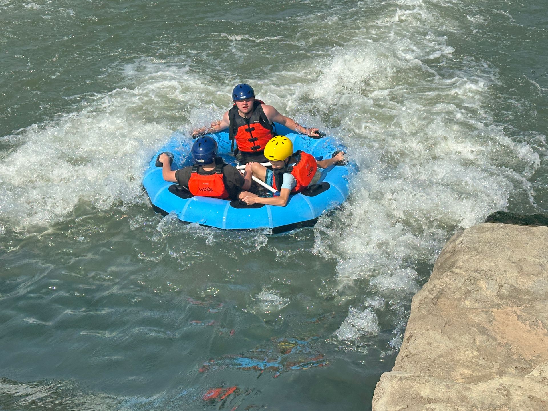 Three people in blue inflatable raft navigating river rapids; wearing helmets and life vests.