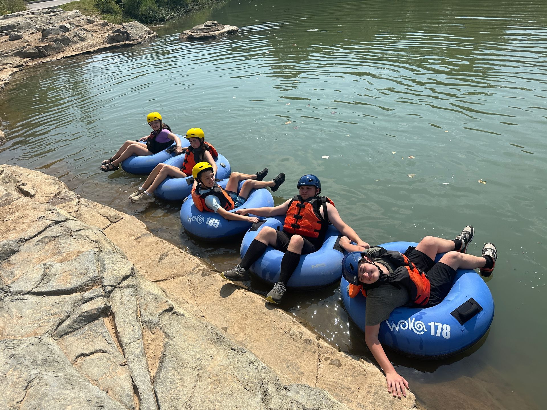 Group of people tubing down a river. They wear life vests and helmets, resting on tubes near a rocky bank.