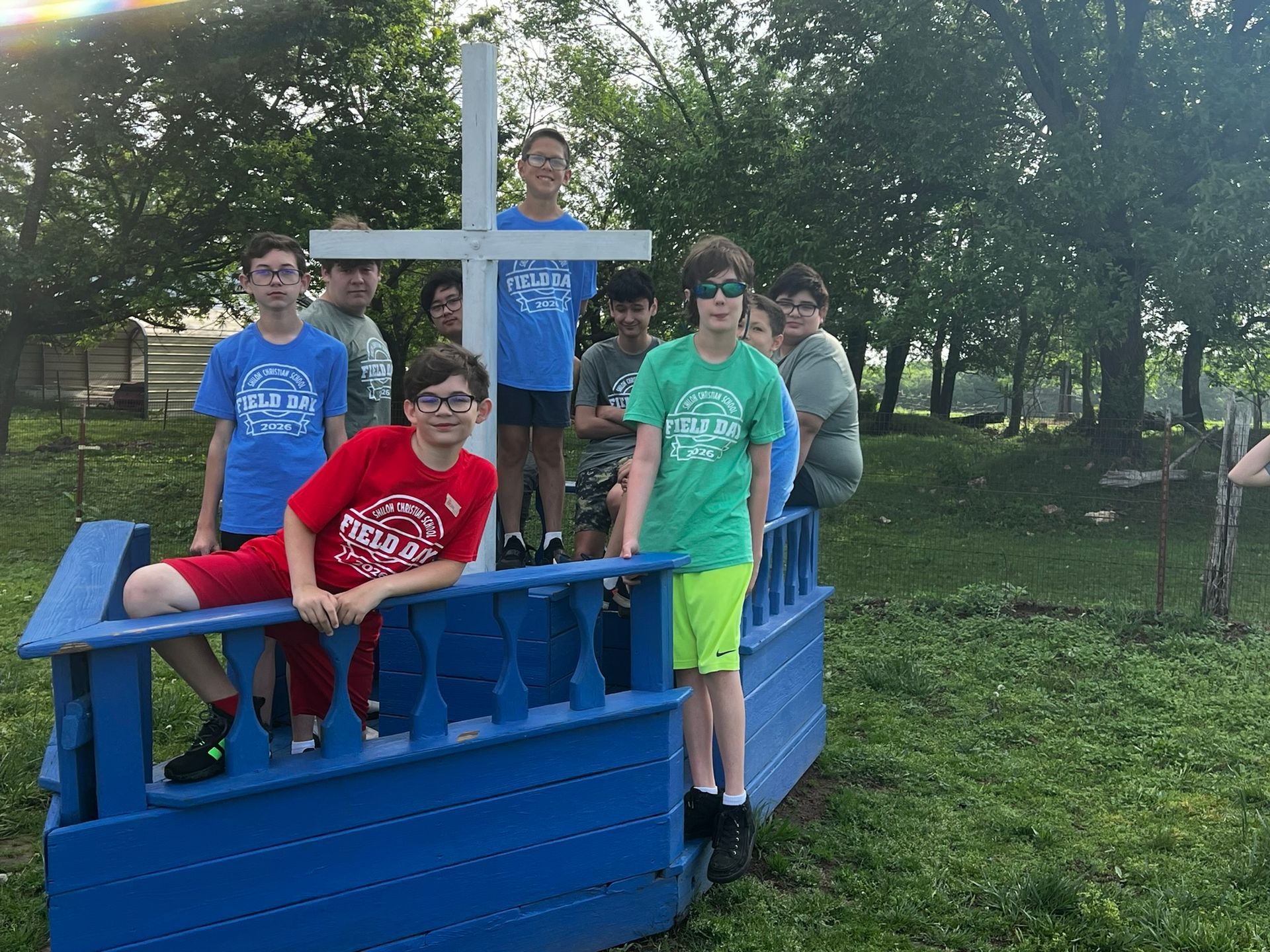 Group of kids posing on a blue wooden boat prop in a grassy park with trees.