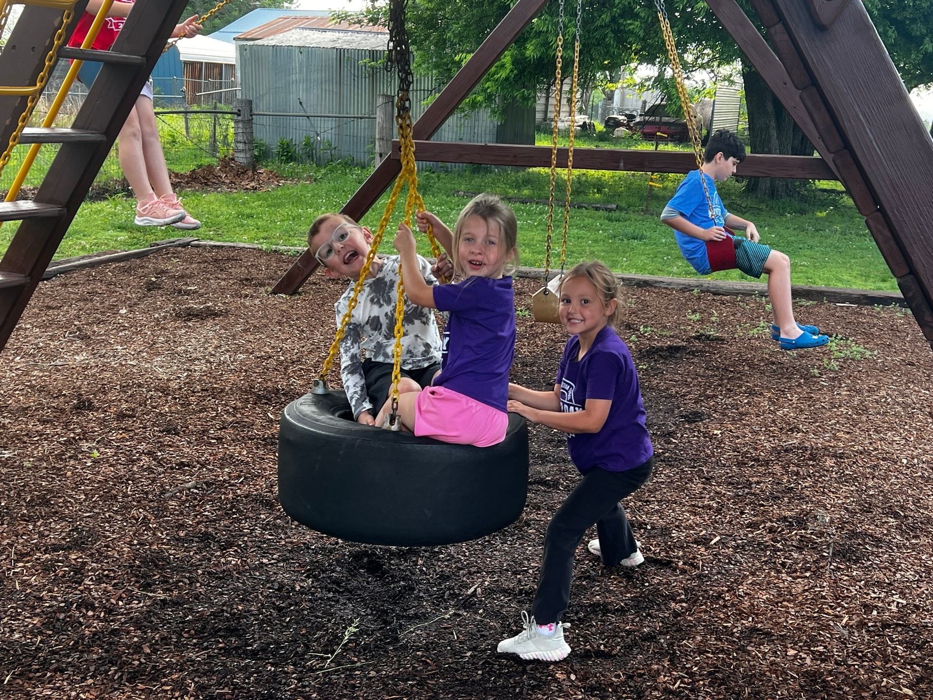 Children playing on a tire swing in a playground, with one girl pushing another on a wood-chip surface.