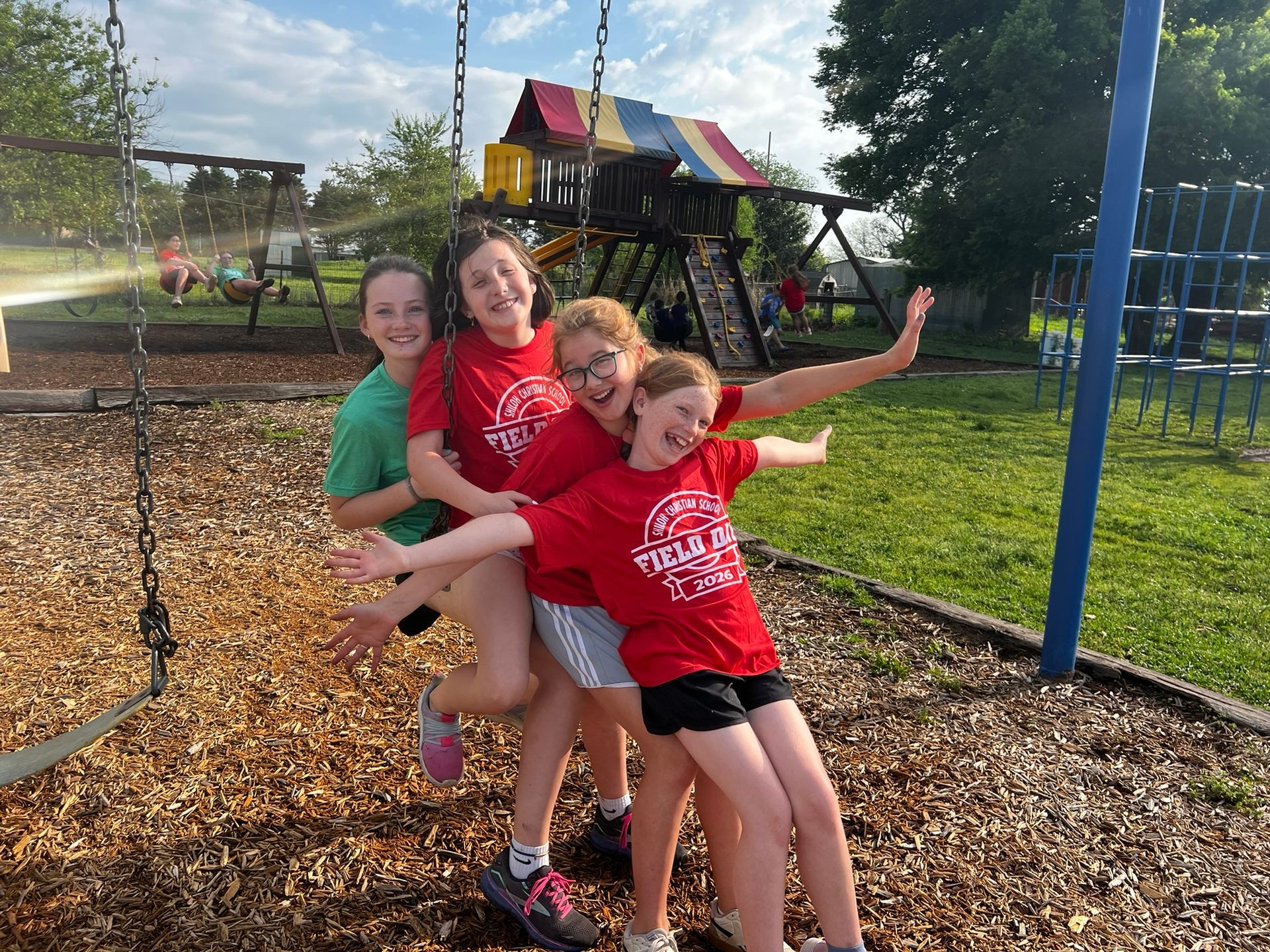 Four children smiling on a playground swing, with a slide and trees in the background.