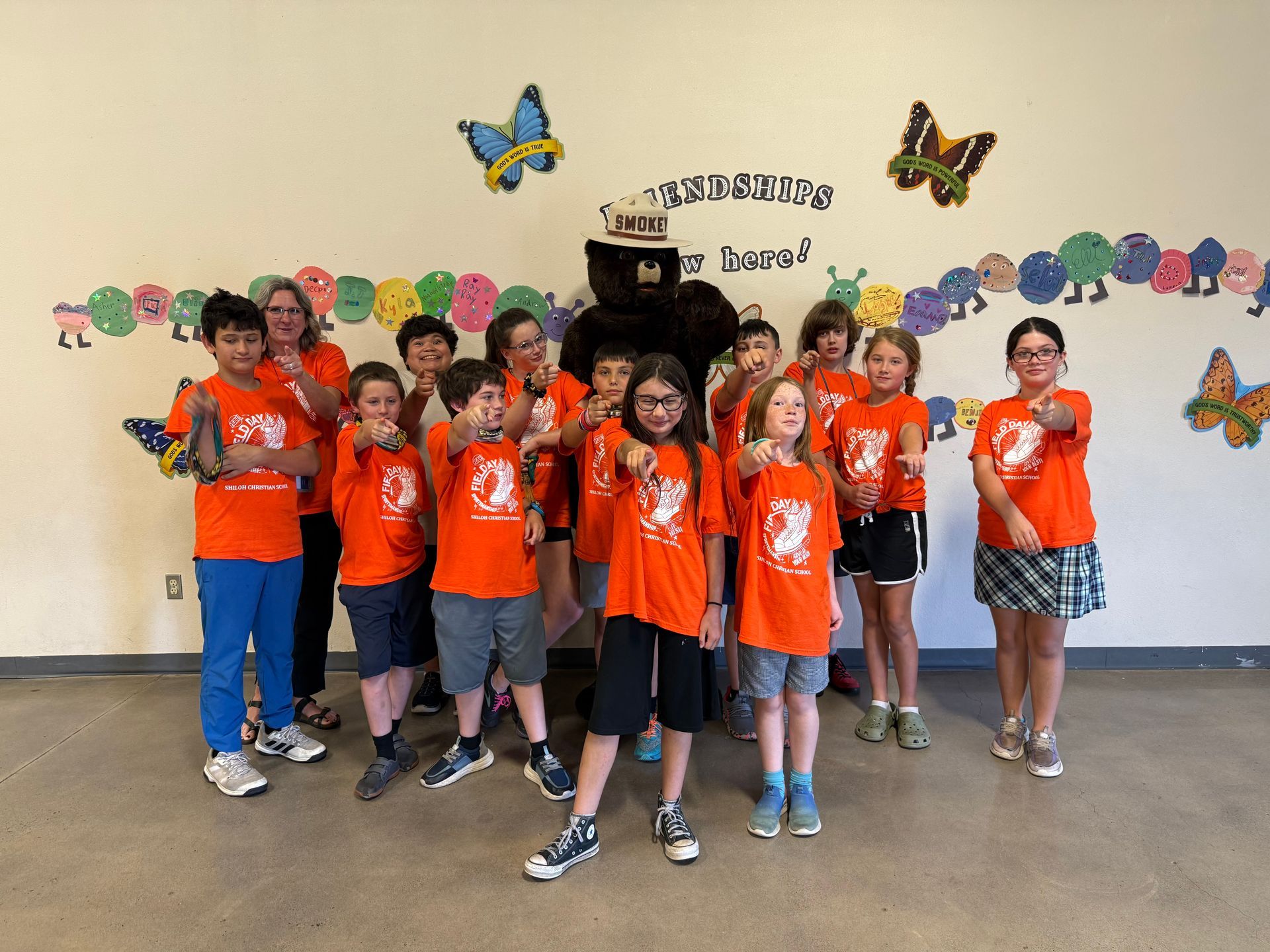 Group of children in orange t-shirts with a mascot, pointing at the camera in a room with colorful decorations.