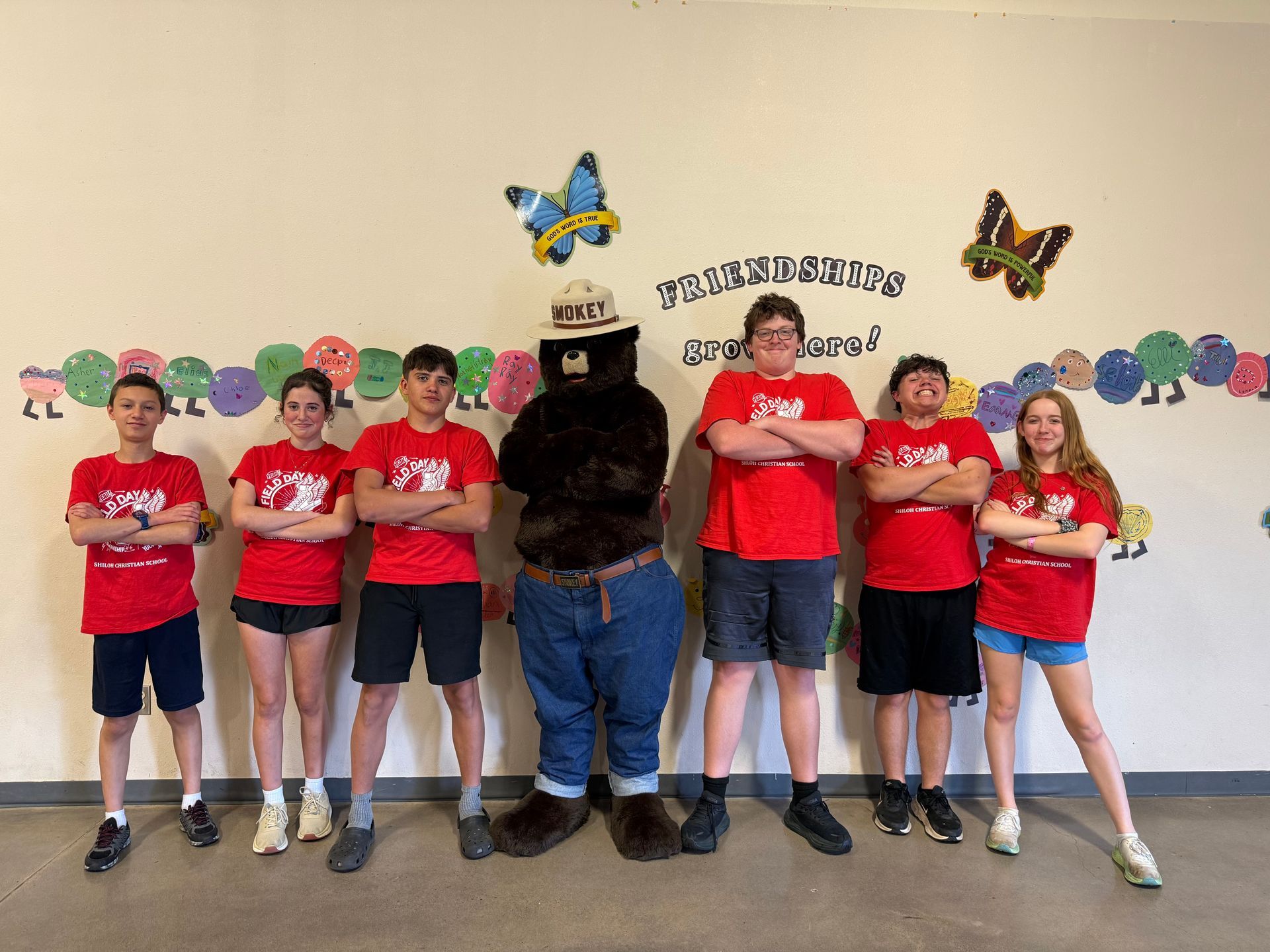 Smokey Bear with a group of children in red shirts posing with arms crossed in front of a colorful wall.