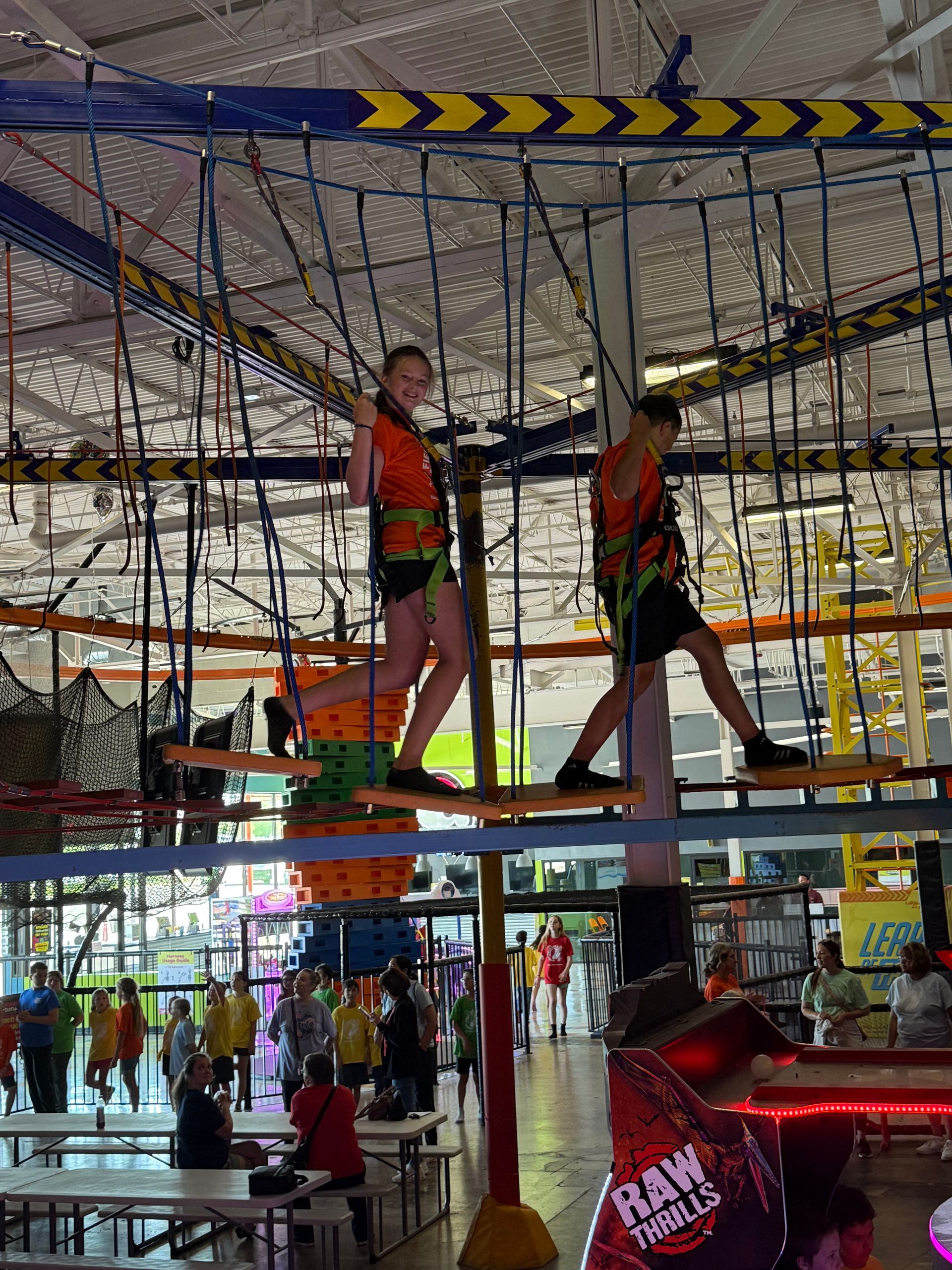 Two people on ropes course, wearing safety harnesses, in a brightly lit indoor recreation center.