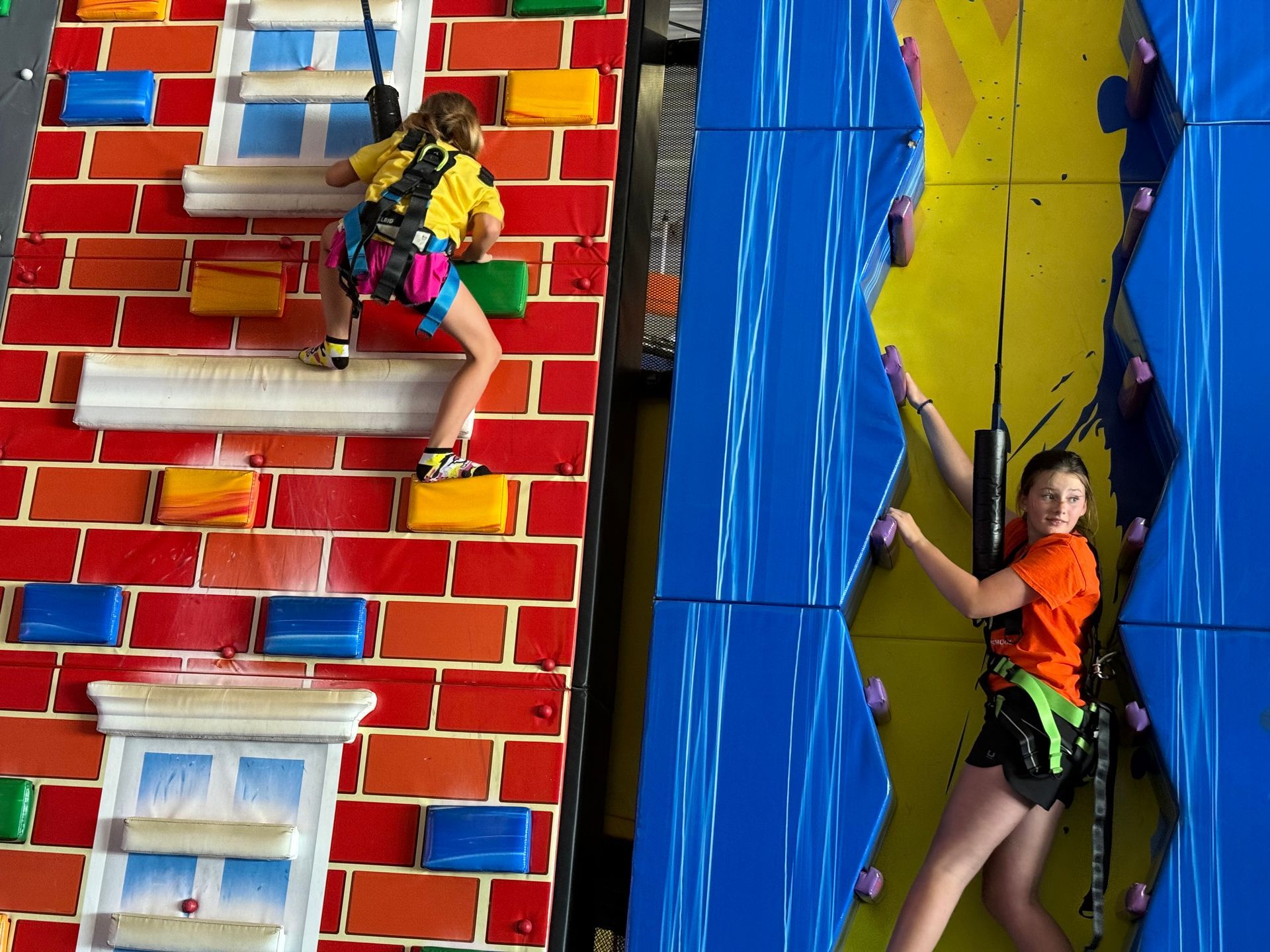 Two people climbing indoor rock walls; brick-like red wall, blue and yellow wall.