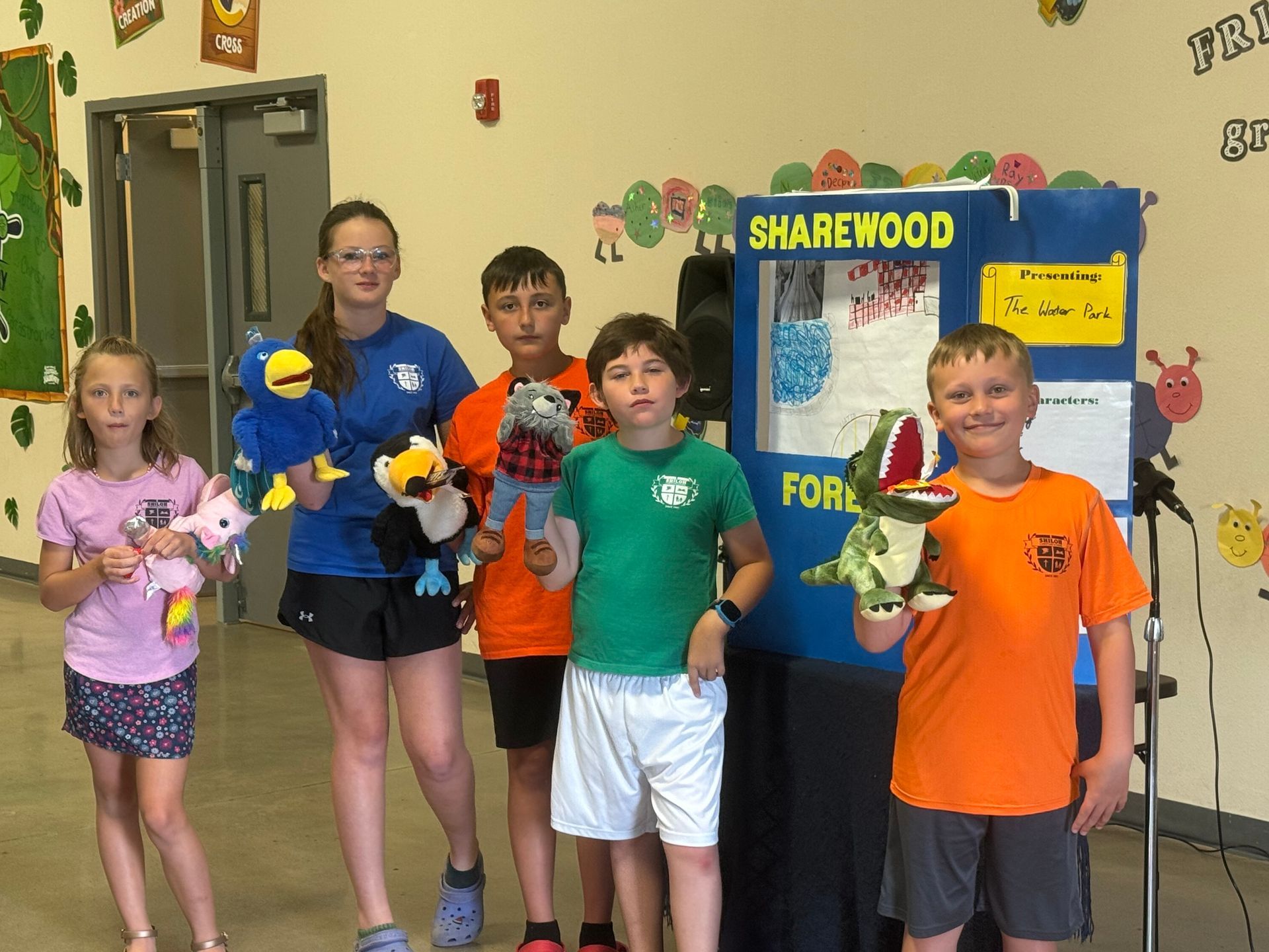 Group of kids with puppets by a puppet stage, in a school hallway.