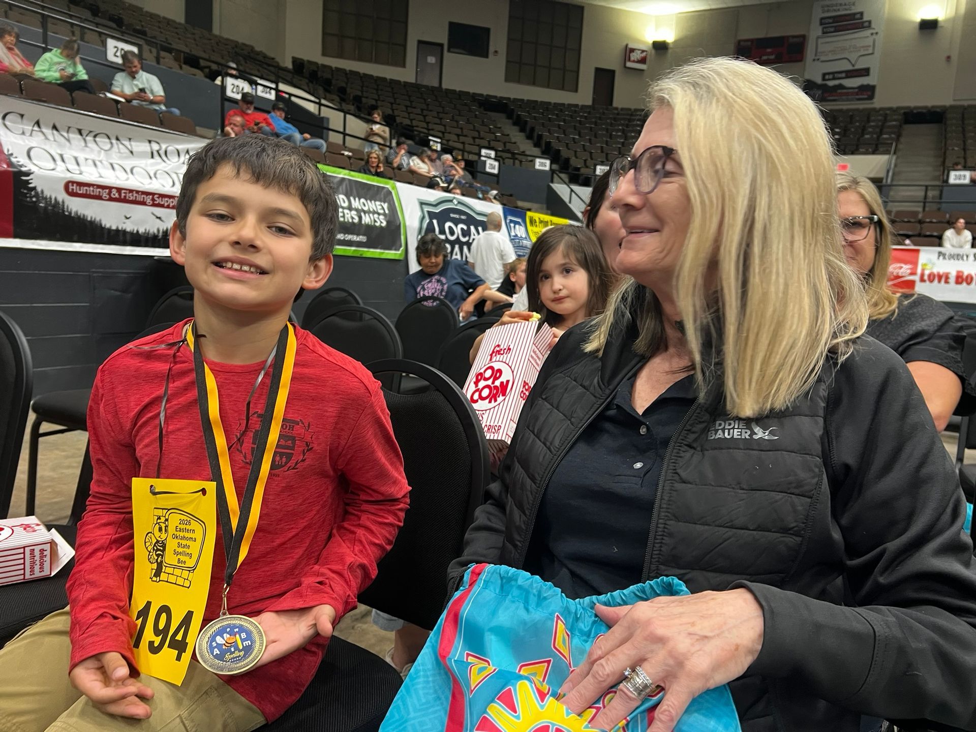 Boy with medal beside a smiling woman in an indoor arena, holding a colorful gift bag.