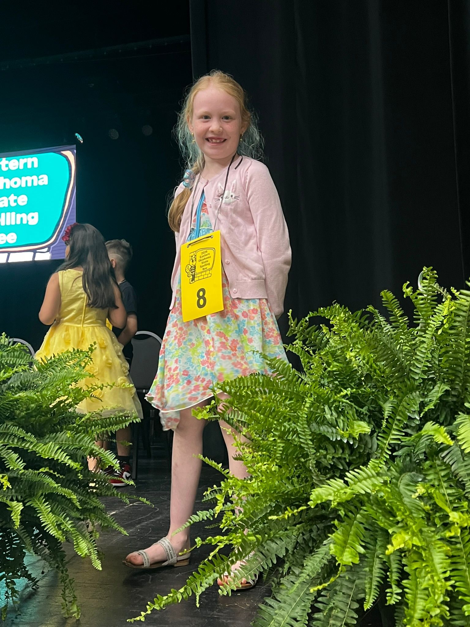 Smiling child in a pink sweater and floral dress stands onstage holding a yellow number tag.