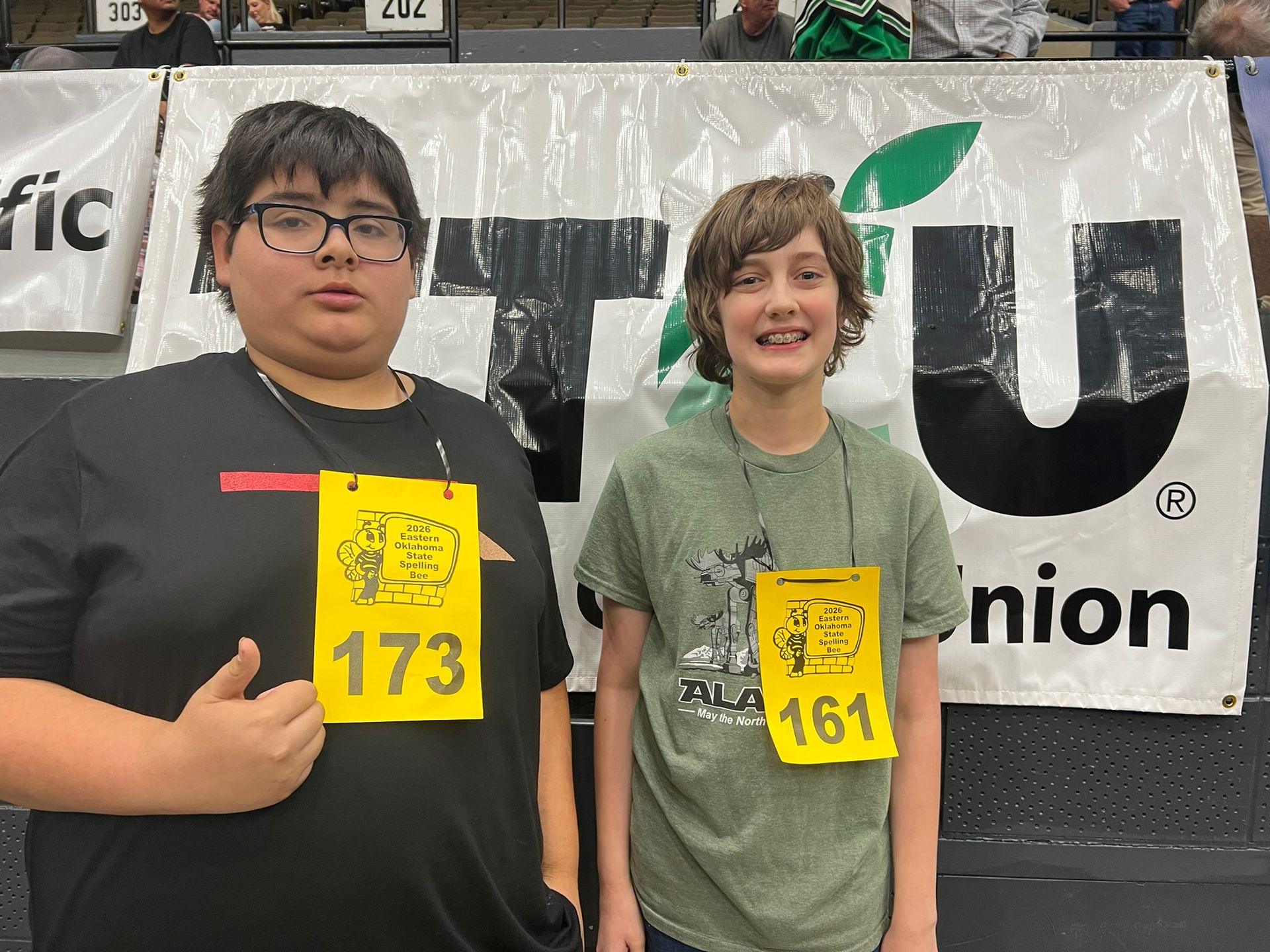 Two runners wearing yellow bibs pose in front of a sponsor banner at an indoor event.