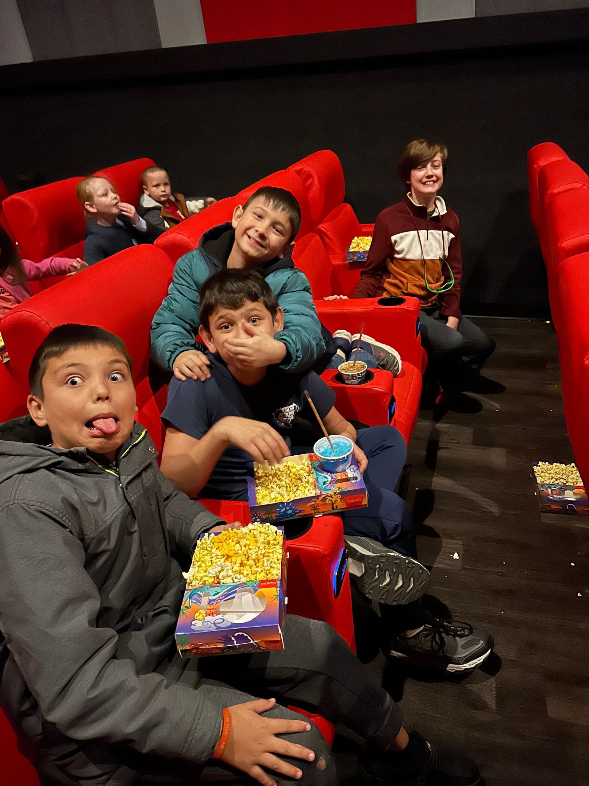 A group of children are sitting in red chairs eating popcorn.