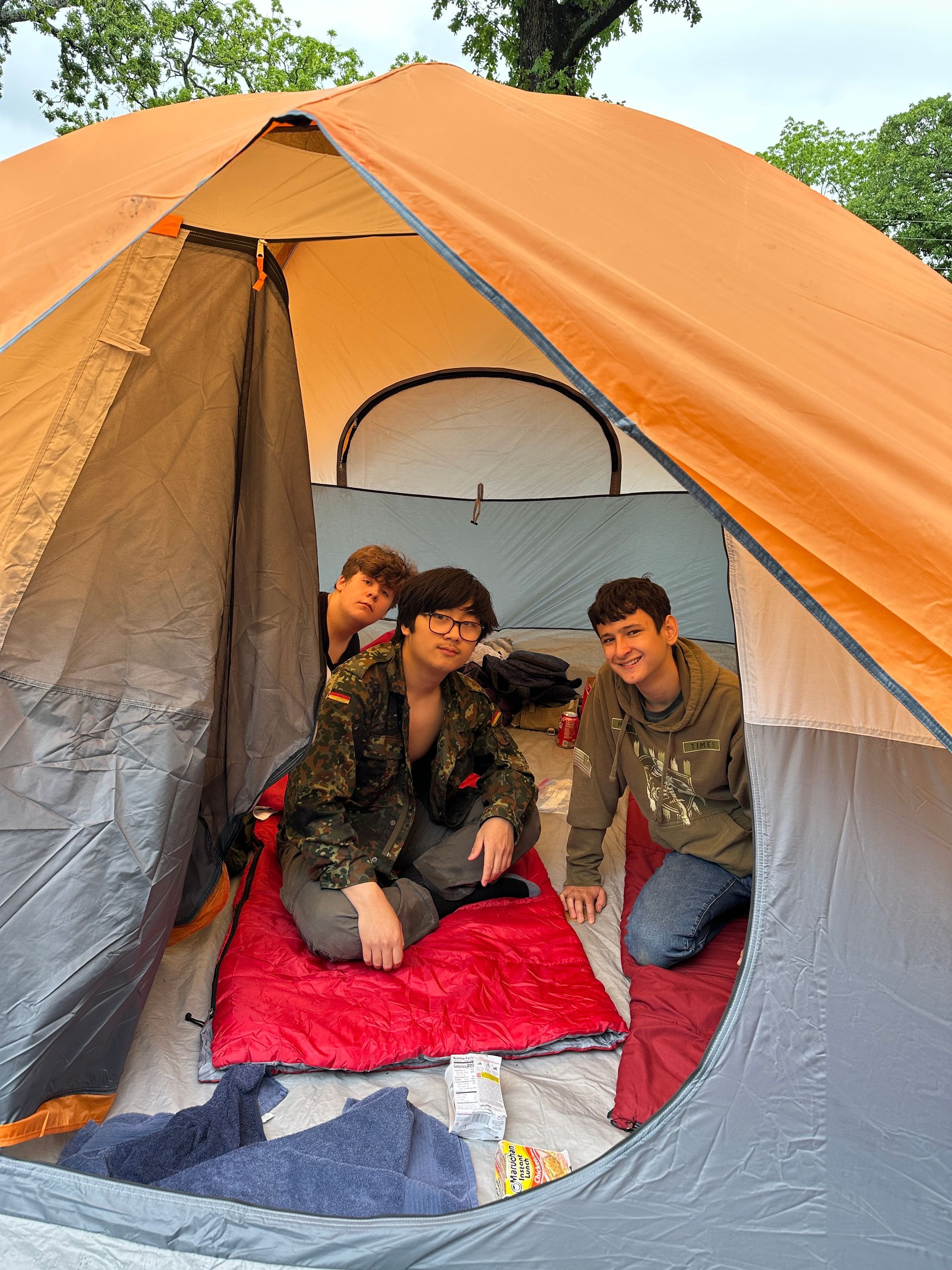 Three people sitting inside an orange camping tent on red and gray blankets.