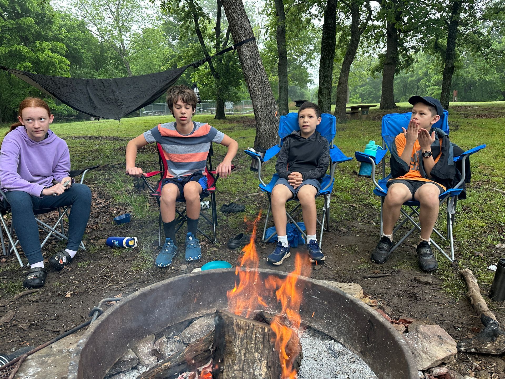 Five children sitting in folding chairs around a campfire in a wooded campsite