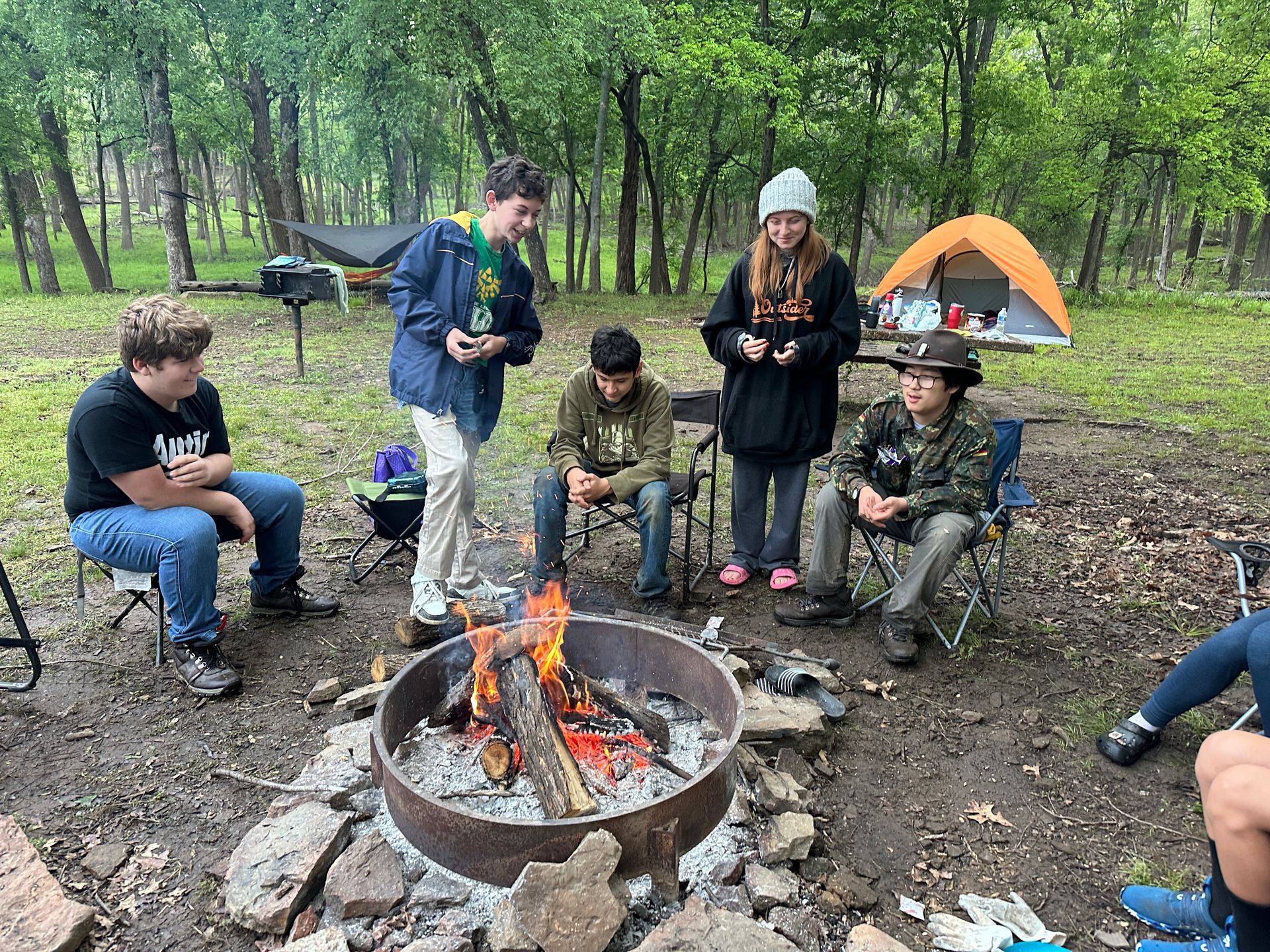 Group of hikers sitting around a campfire in a forest, with a small tent in the background.