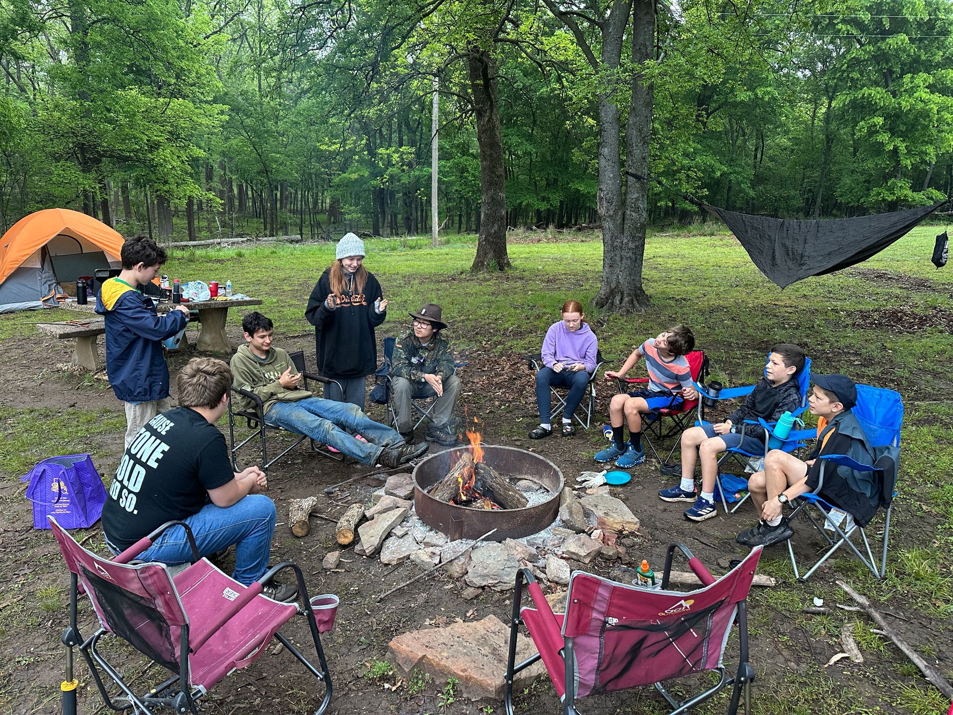 People sitting around a campfire in a wooded campsite with tents nearby
