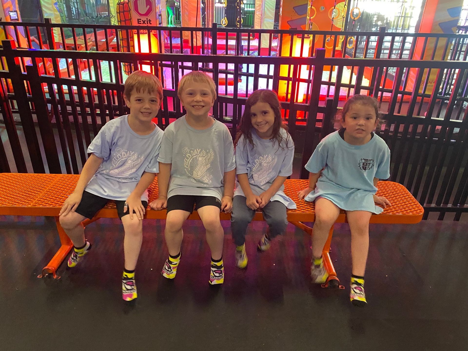 Four children in blue shirts sitting on an orange bench at an indoor playground.