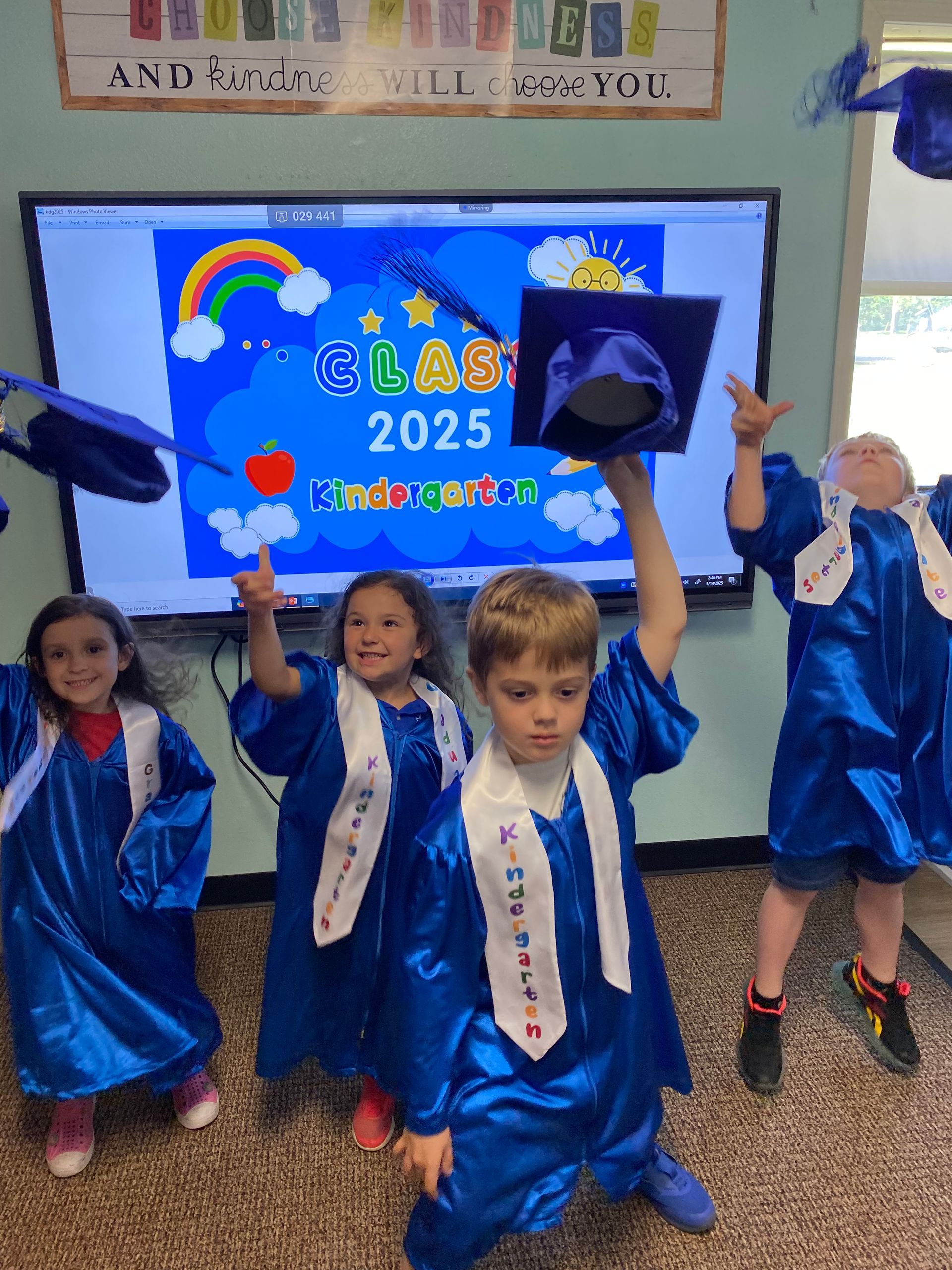 Kindergarten graduation: Children in blue gowns throwing caps, celebrating, and smiling in front of a screen.