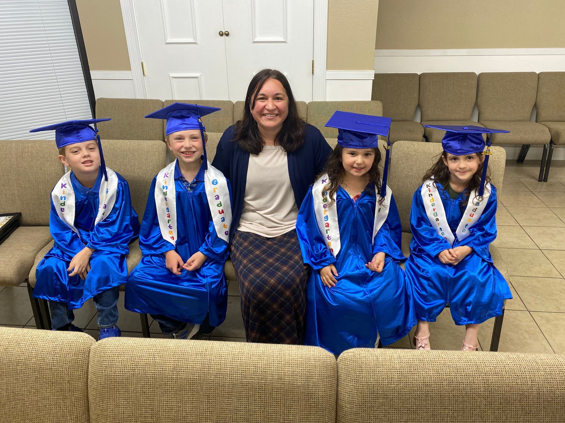 A teacher and five children in blue graduation gowns smile on a tan couch.