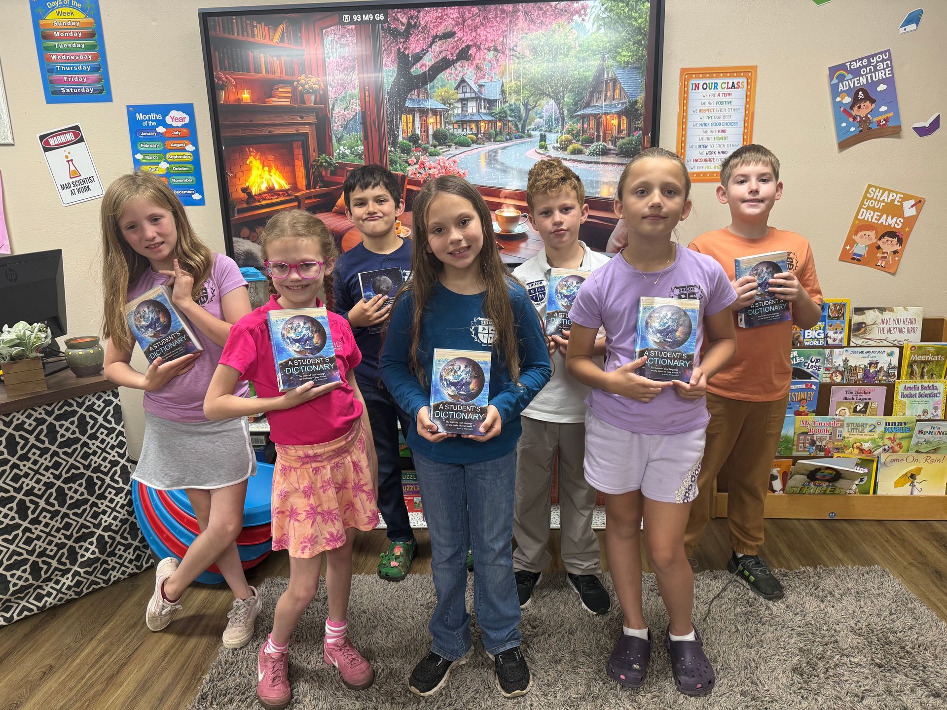 Children holding books in a colorful classroom, standing in front of a winter fireplace scene.