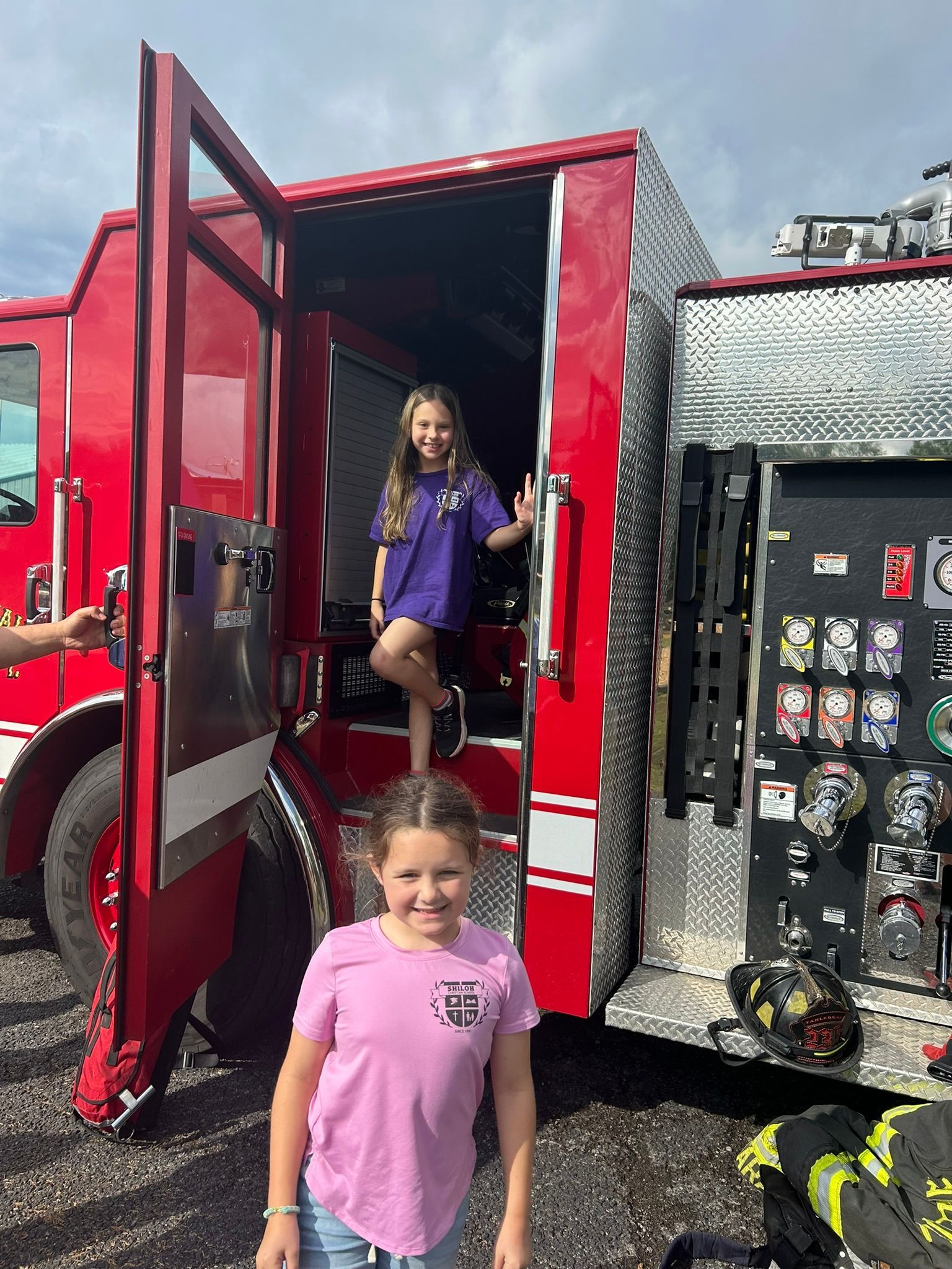 Two girls pose near a red fire truck with an open door. One girl is inside, the other stands in front.