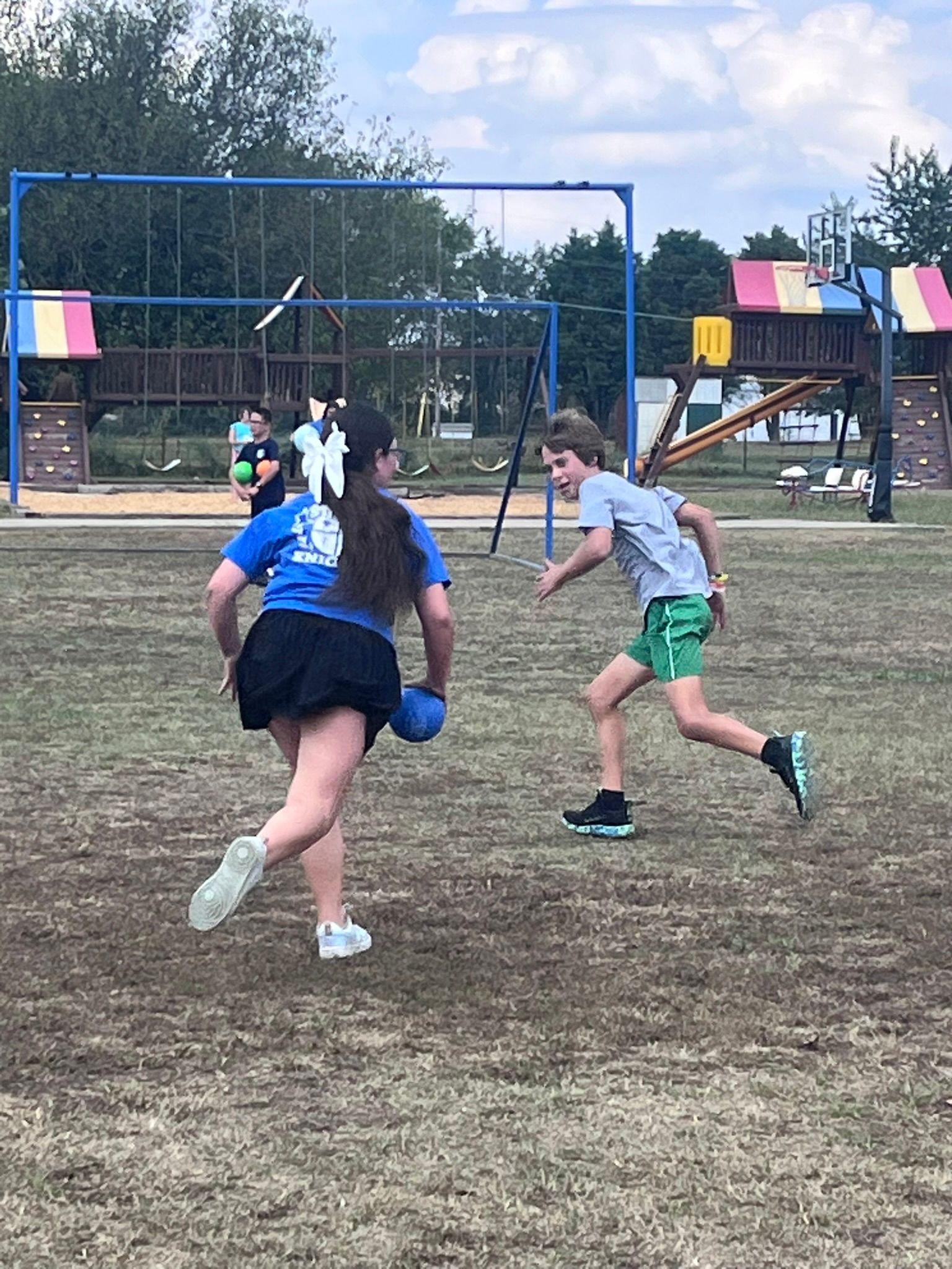 Two children playing dodgeball on a grassy field near a playground.