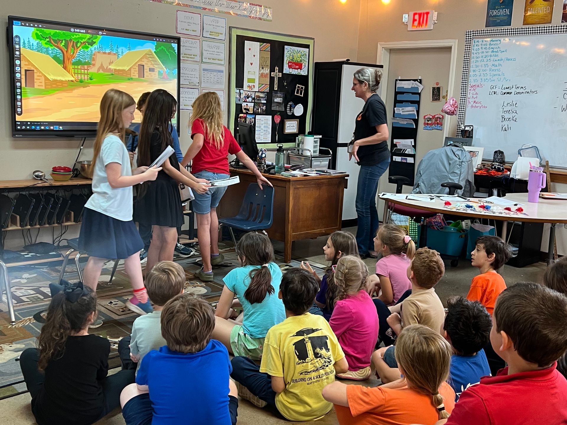Children in a classroom with a teacher. Some present to others. One screen shows a cartoon.