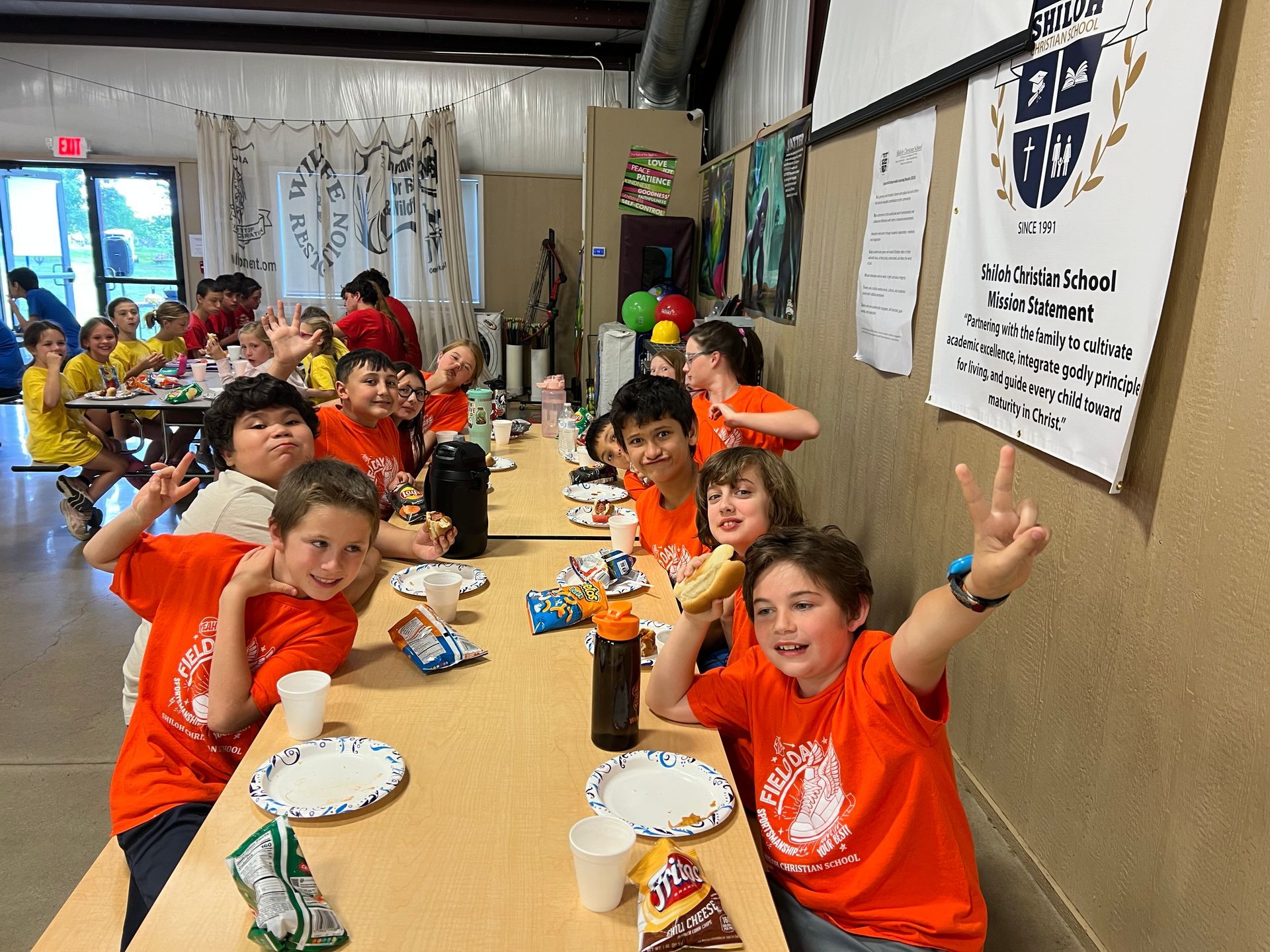 Kids in orange shirts at a long table, smiling and making peace signs indoors.