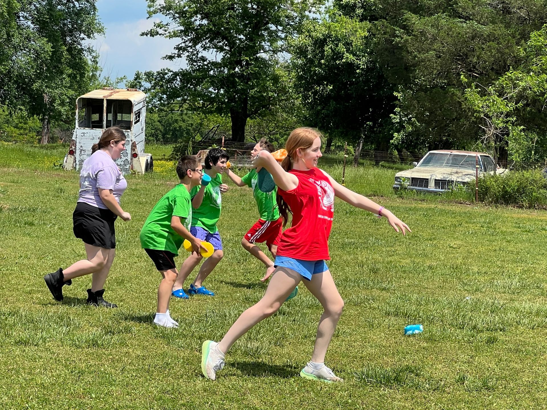 Children playing a throwing game in a grassy field with a wrecked car and a trailer.