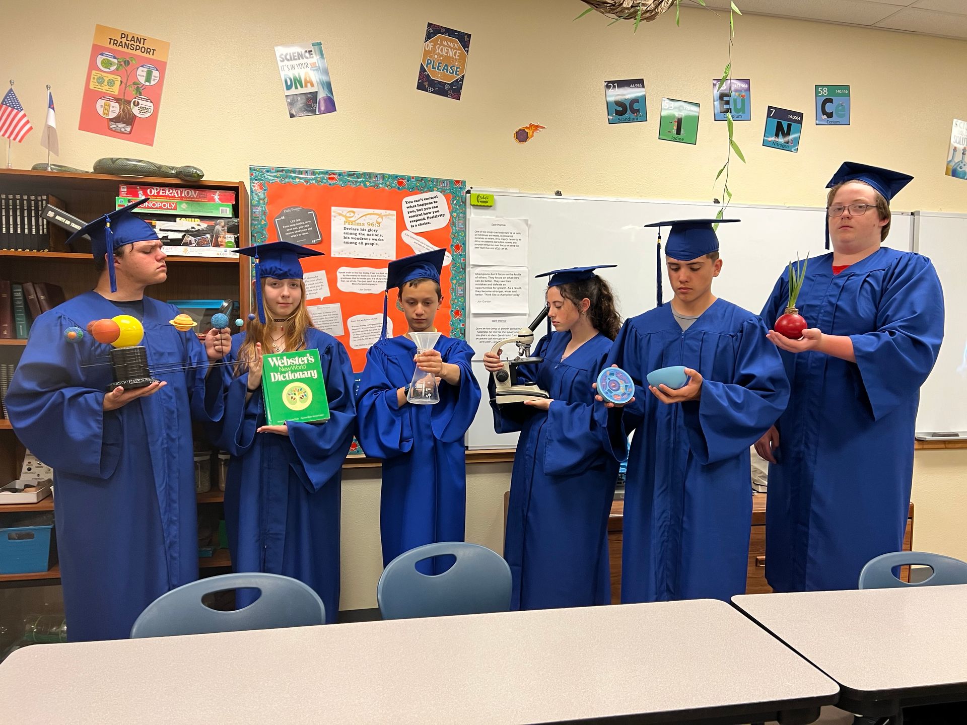 Seven people in blue graduation gowns and caps stand in a classroom, holding items.