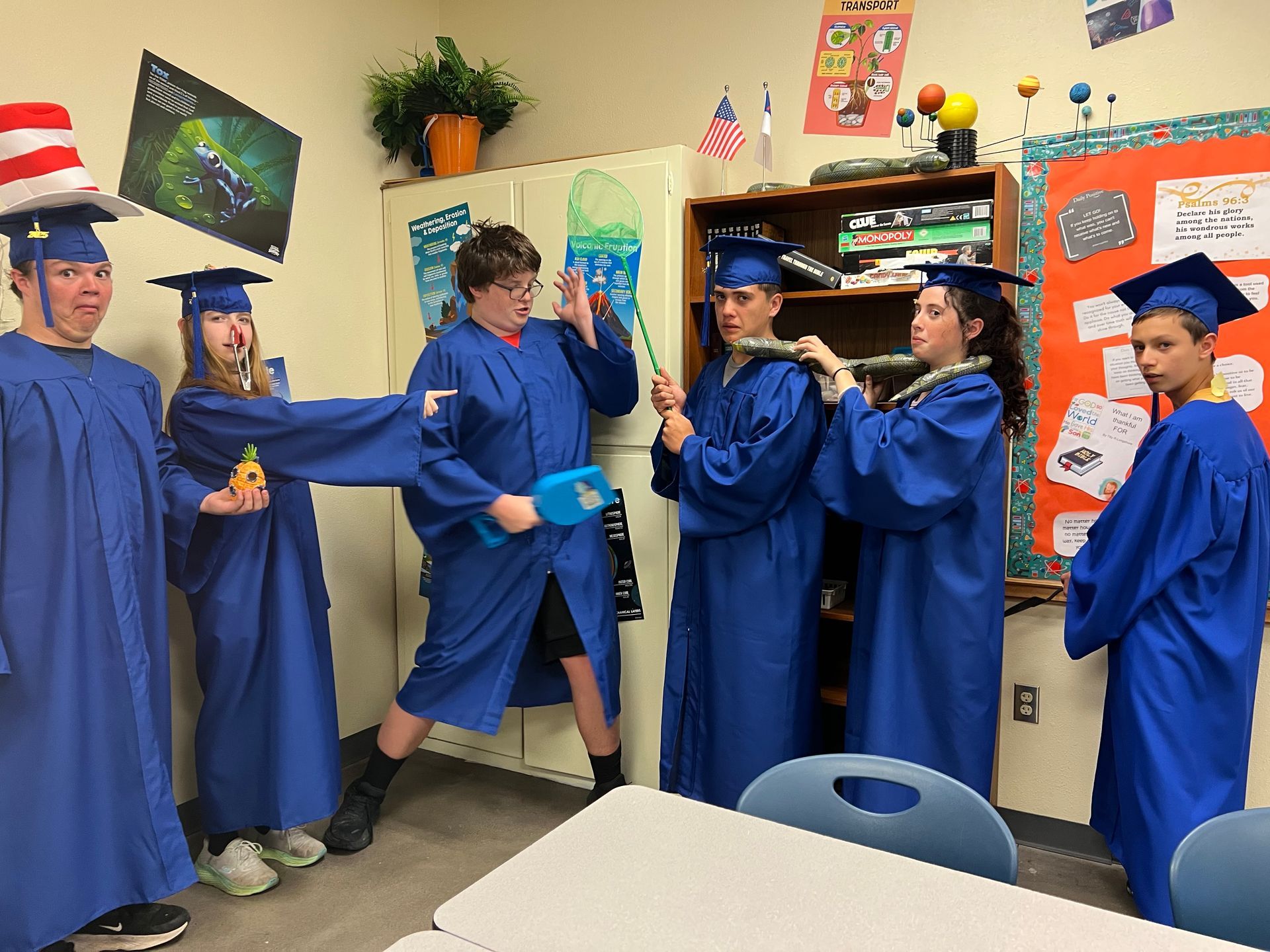 Six students in blue graduation gowns and hats, clowning around in a classroom. One wears a Cat in the Hat hat.