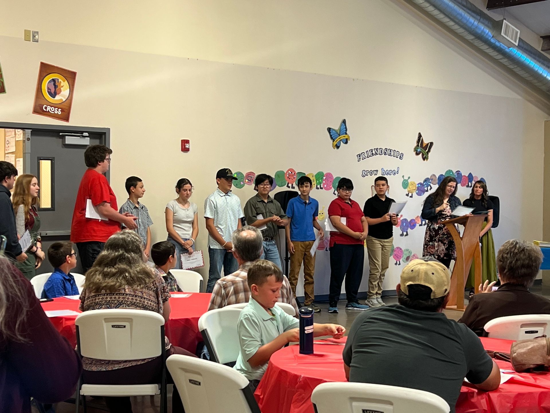 A group of students presenting at an event. People sitting at red tables, indoors with decorations.