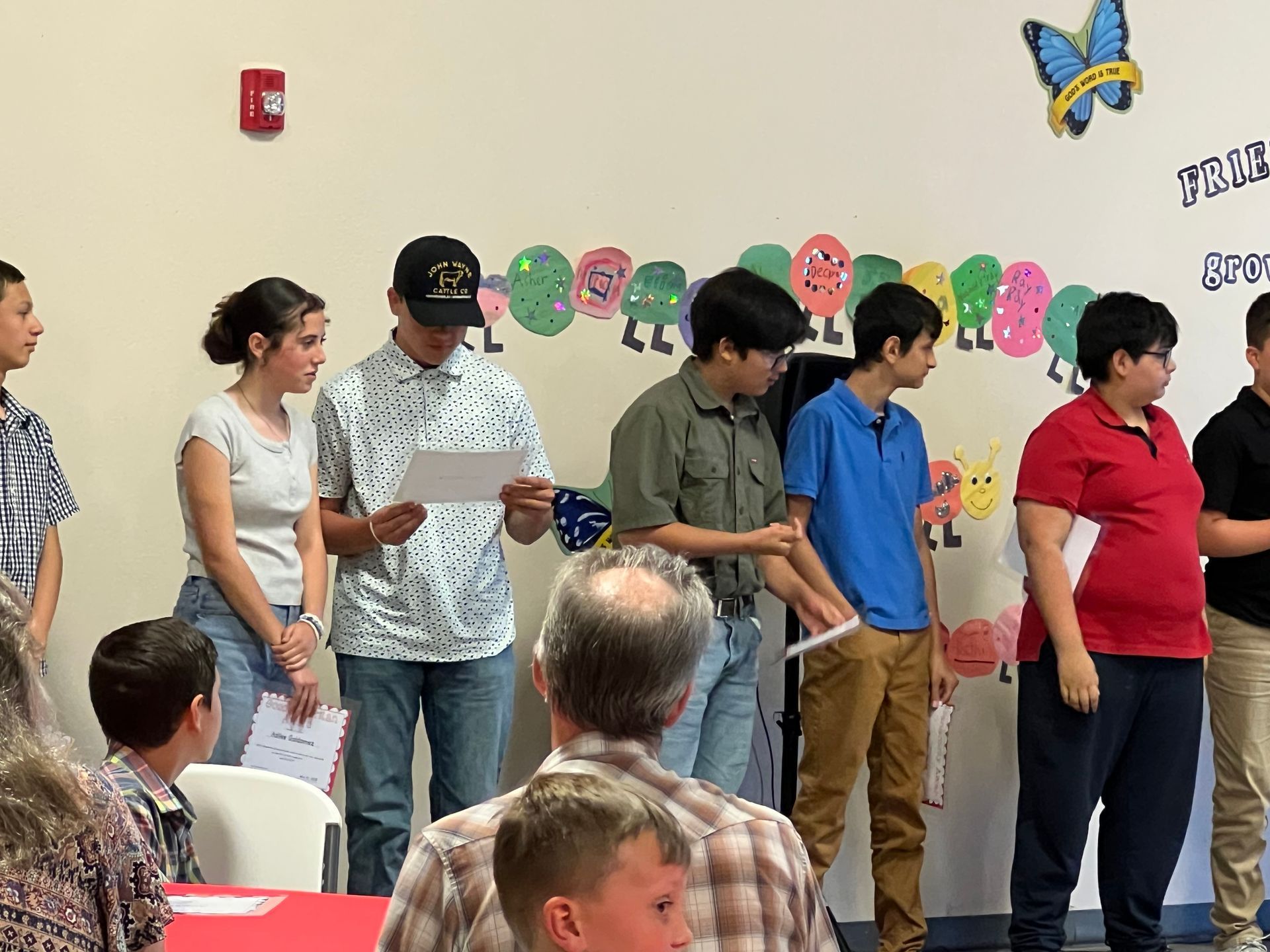 Group of teens stand holding papers, near a decorated wall. Some appear to read, inside.