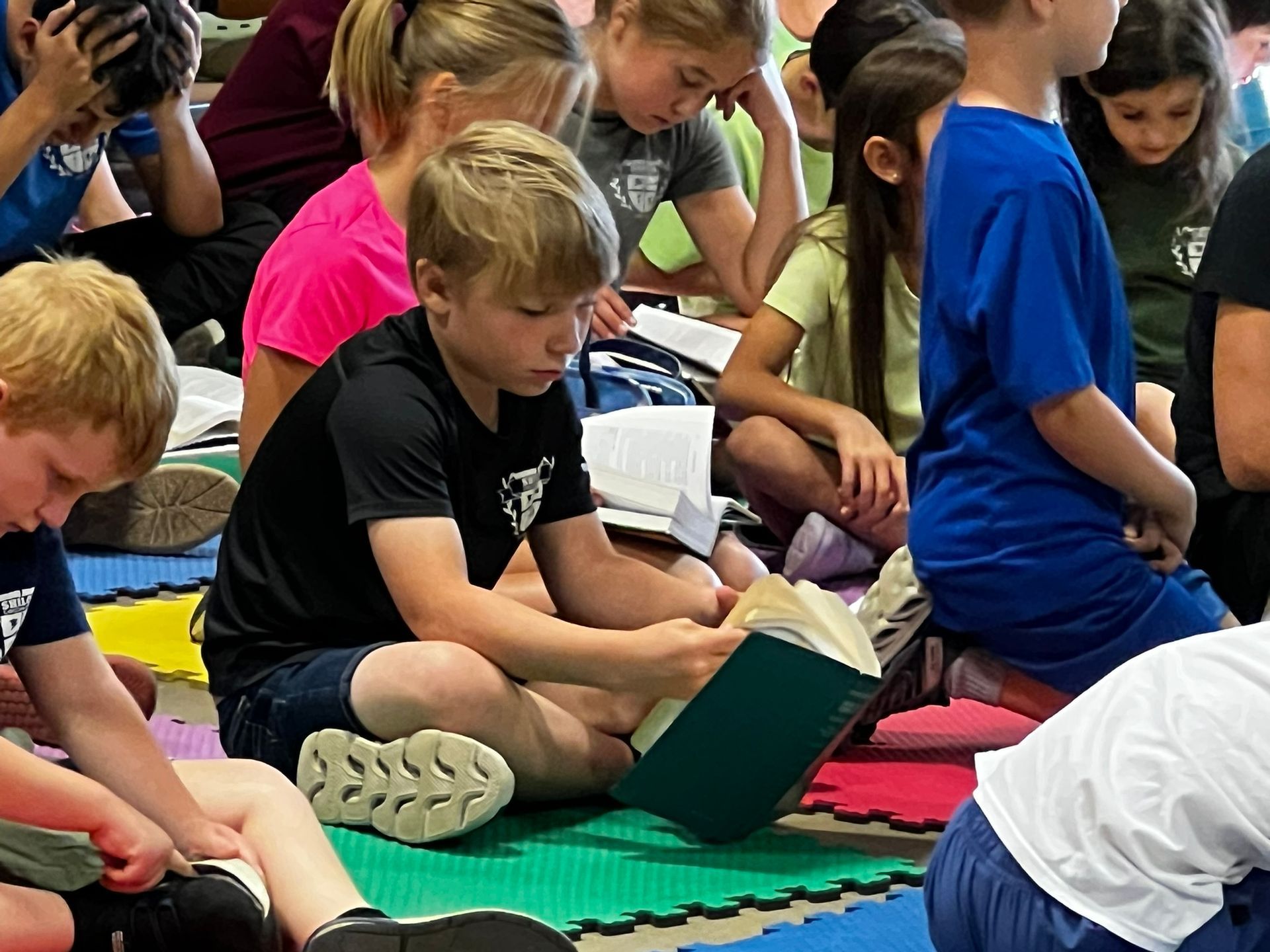 Children in casual clothes reading books while sitting on the floor, likely in a classroom.