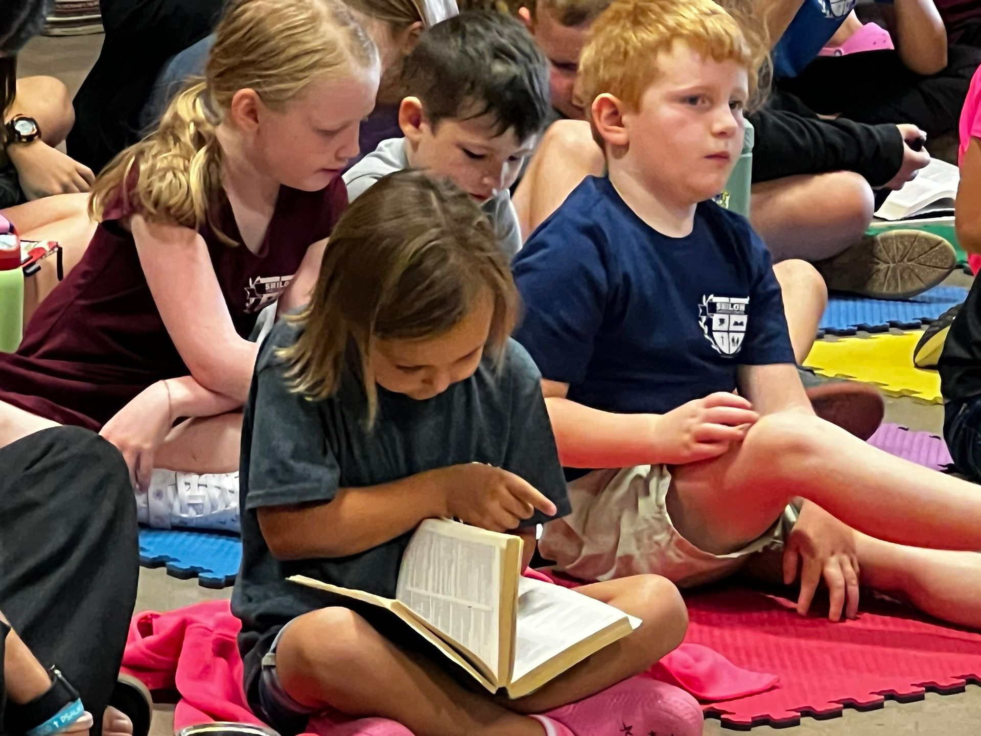 Children reading books, sitting on the floor. One points to a page, others appear focused.