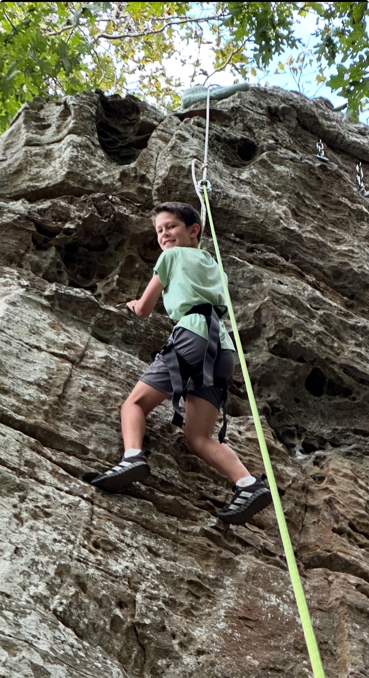 Boy in green shirt climbs a rocky cliff, secured by a rope and harness. Smiling, outdoors.