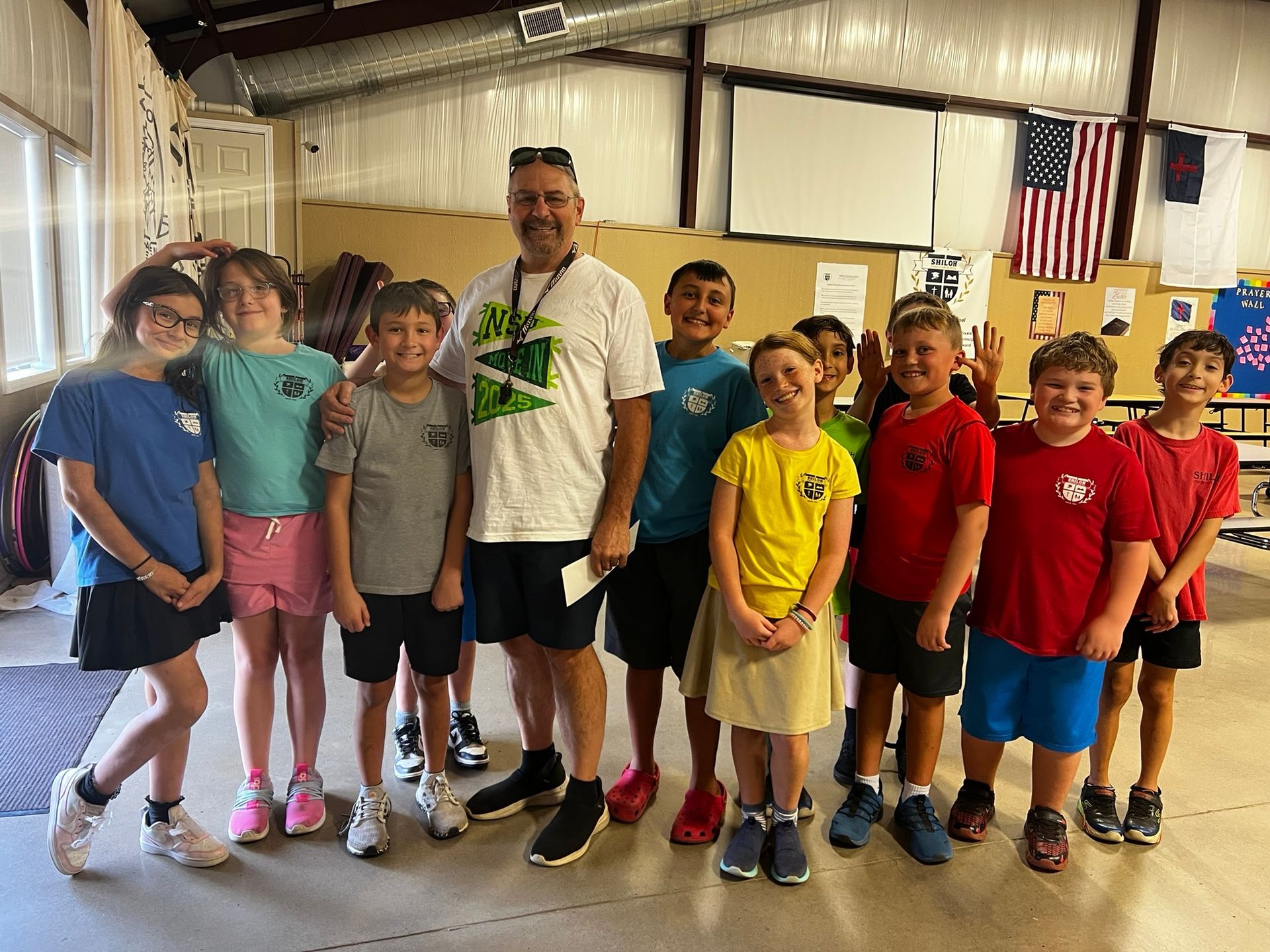 Group of children and an adult smiling, standing indoors; American flag in background.
