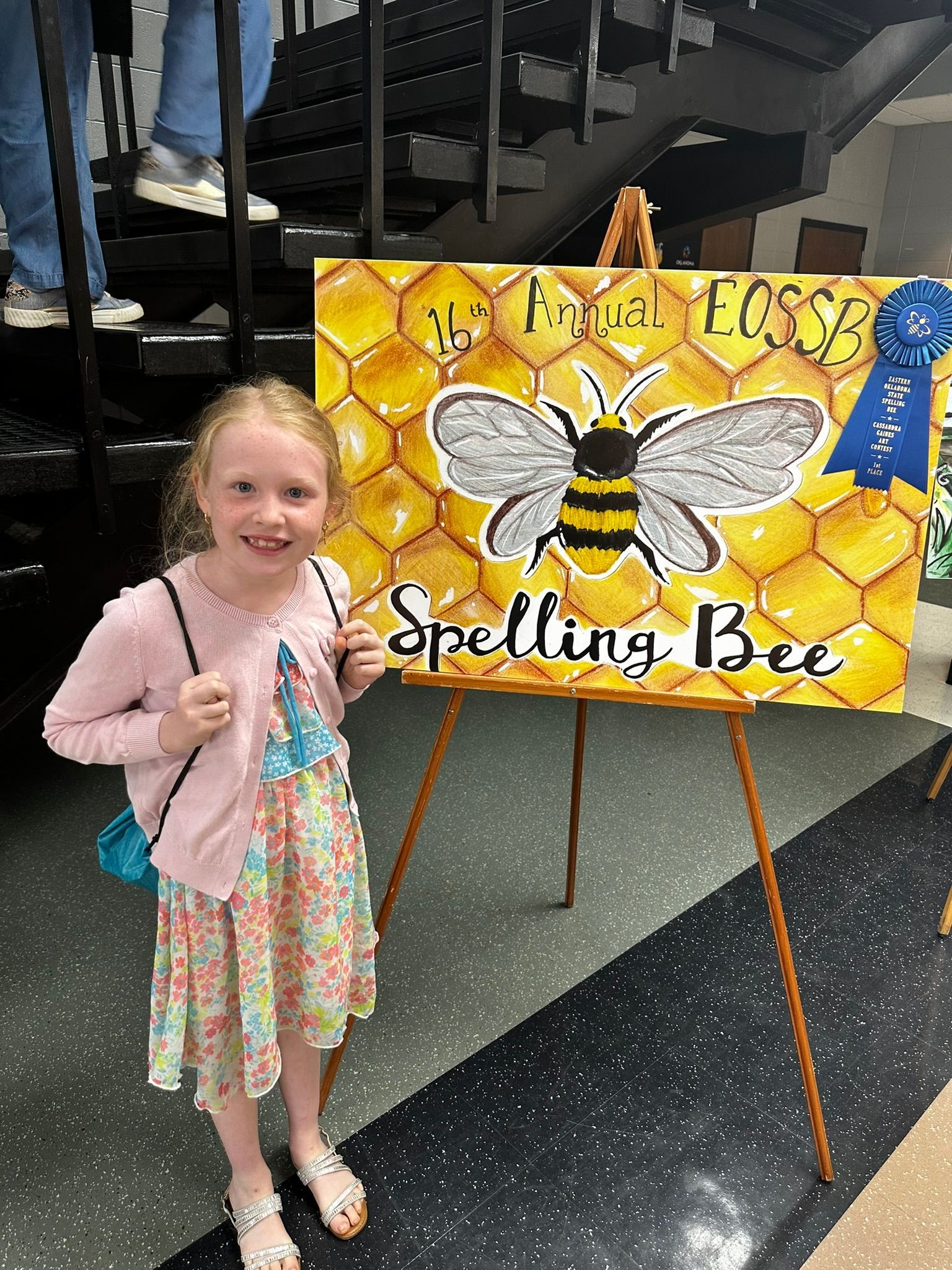 Young girl standing beside a spelling bee sign with a bee illustration on an easel