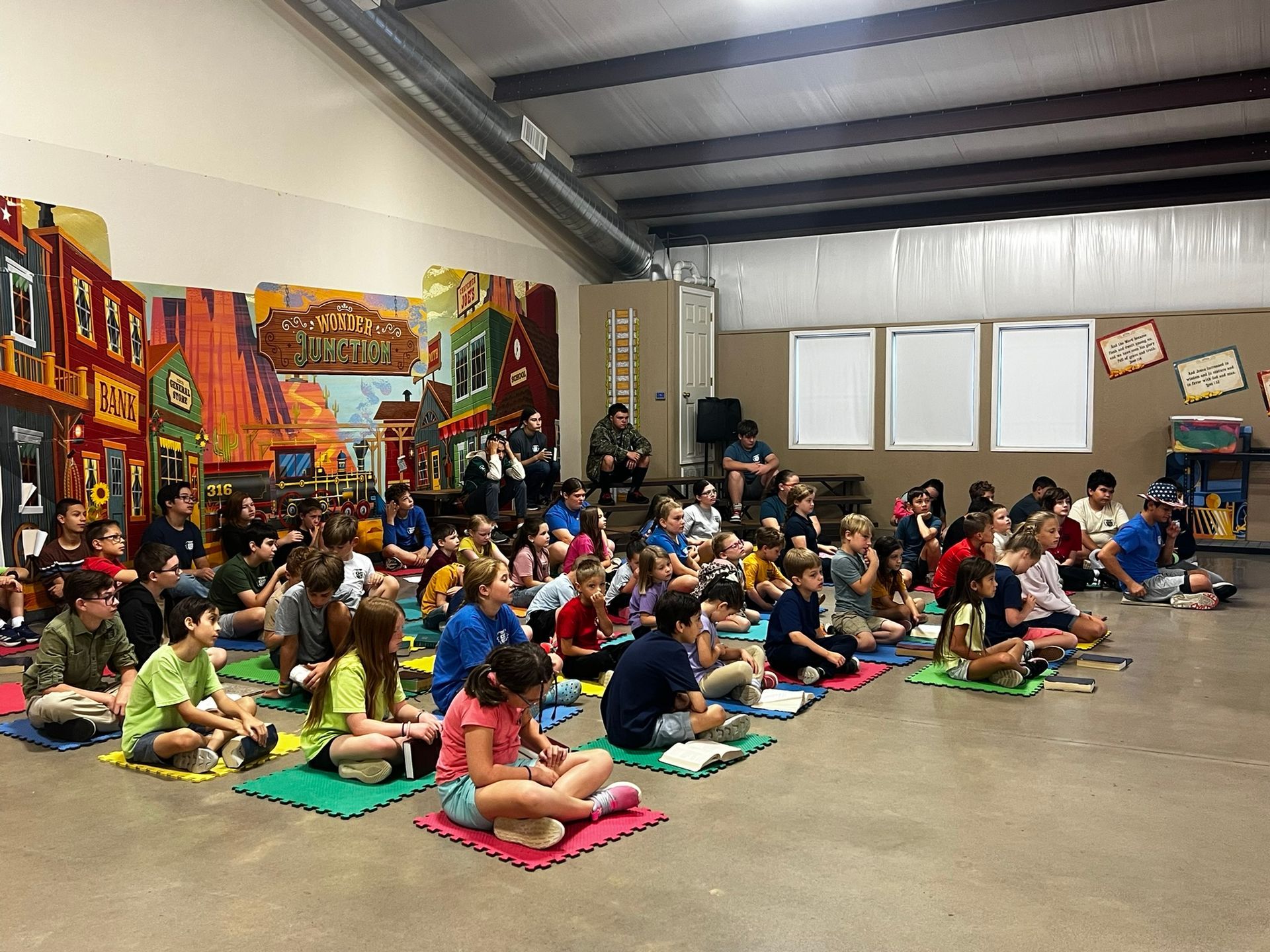 Children seated on mats in a large room listening, colorful mural in the background.