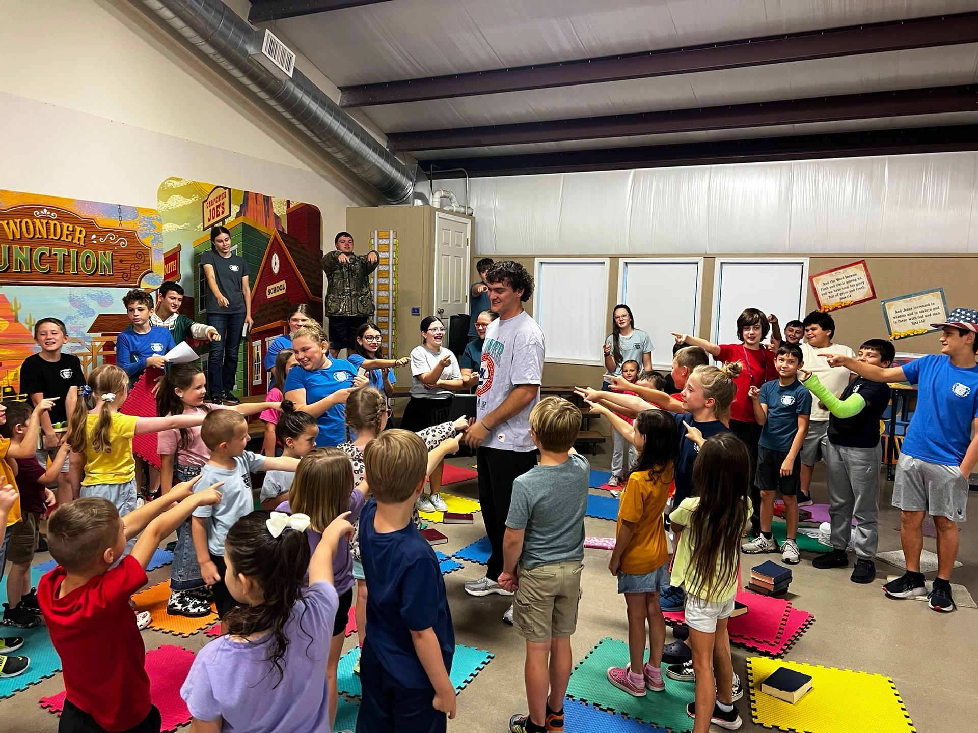 Children pointing at a man in a room with others, colorful mural and mats, possibly a camp or activity.