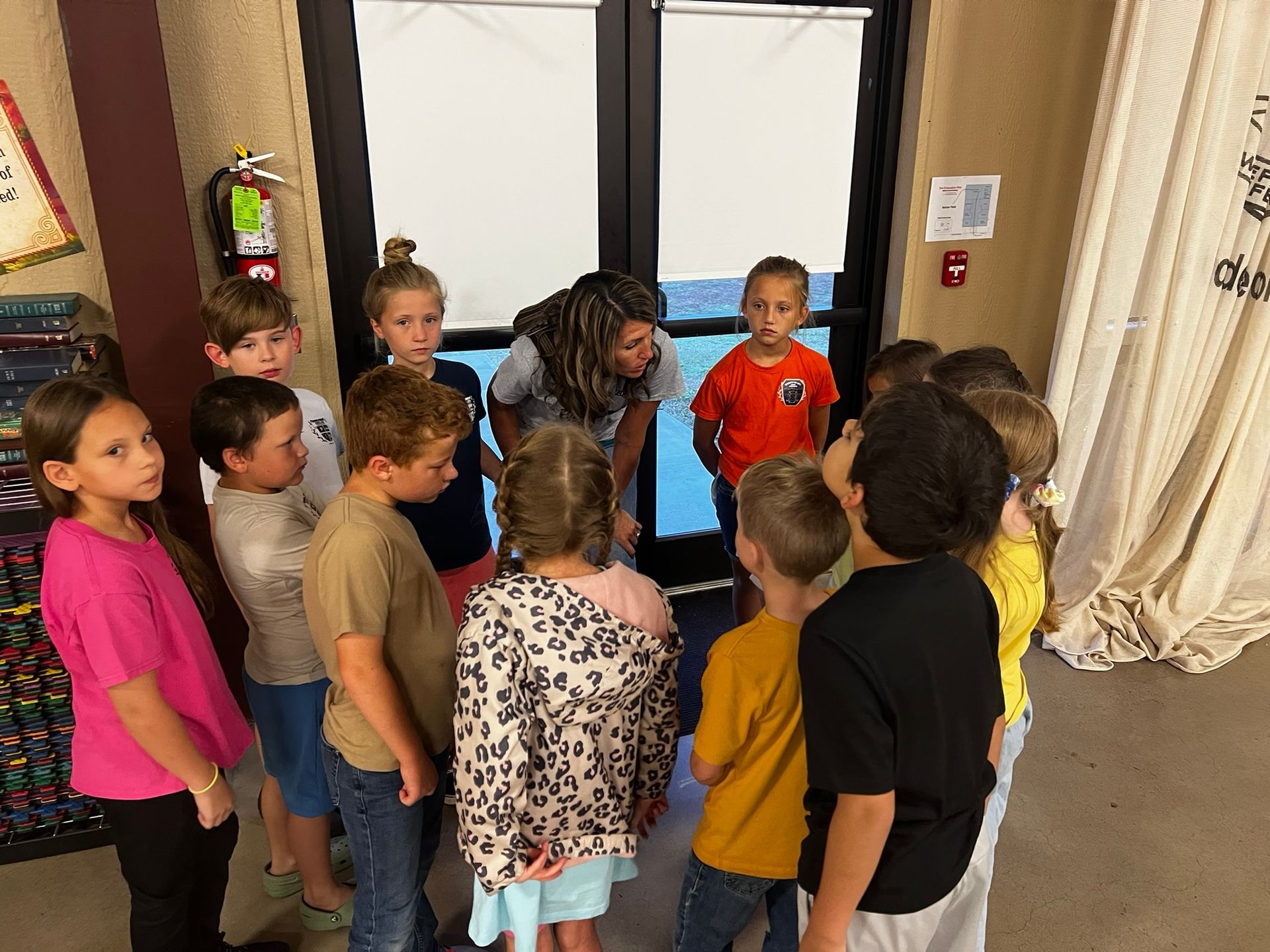 A teacher and a group of children clustered around, focused, indoors near a doorway.
