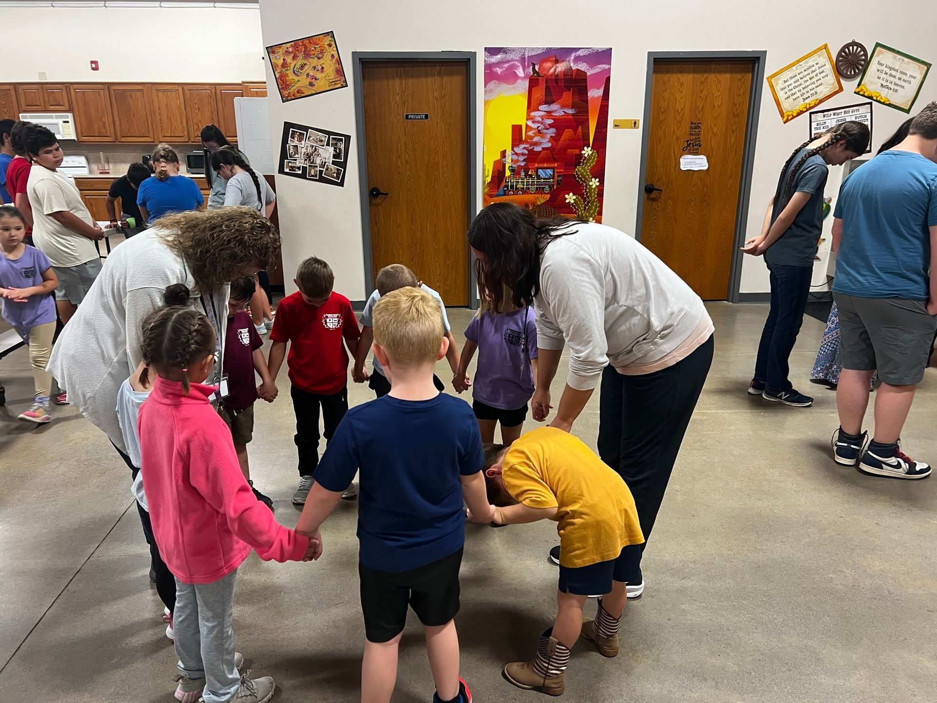 Children and adults in a circle holding hands, playing a game in a room with doors and artwork.