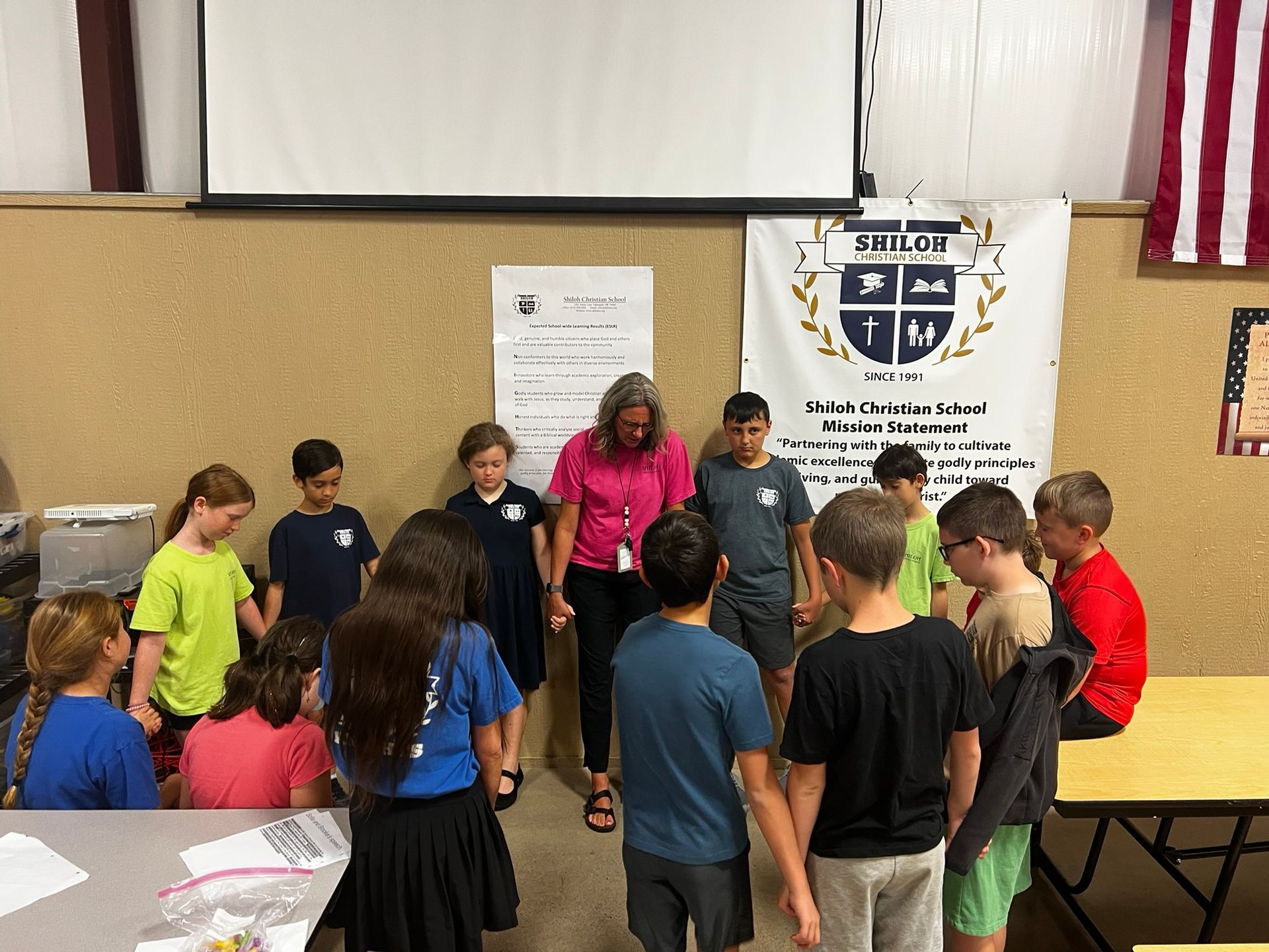 Children and teacher holding hands in a circle, indoors, at a school, with a banner.