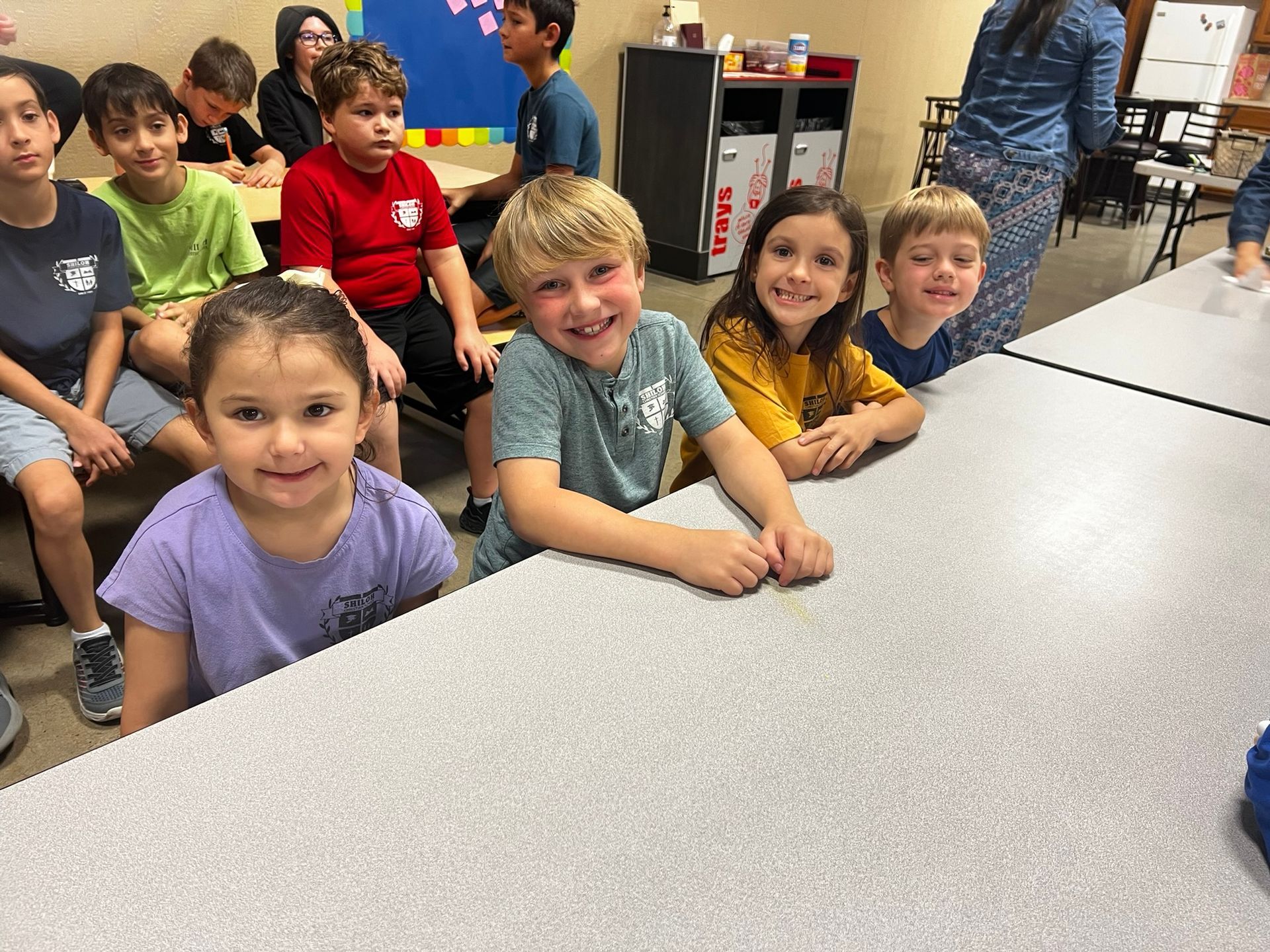 Children smiling at a table. Blonde boy smiles widely. Other children in various colored shirts. Indoors.