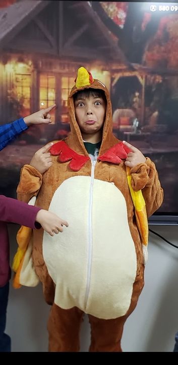 Person in a turkey costume makes a face. Two hands point at the costume. Background has a fall scene.