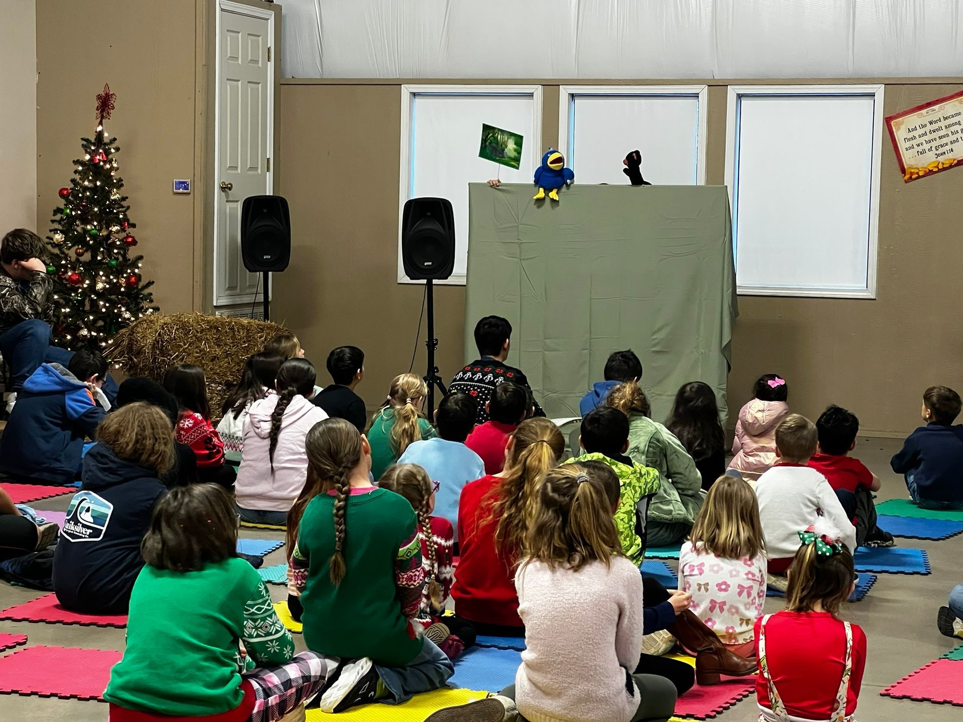 Children watch a puppet show indoors; green backdrop, speakers, Christmas tree.