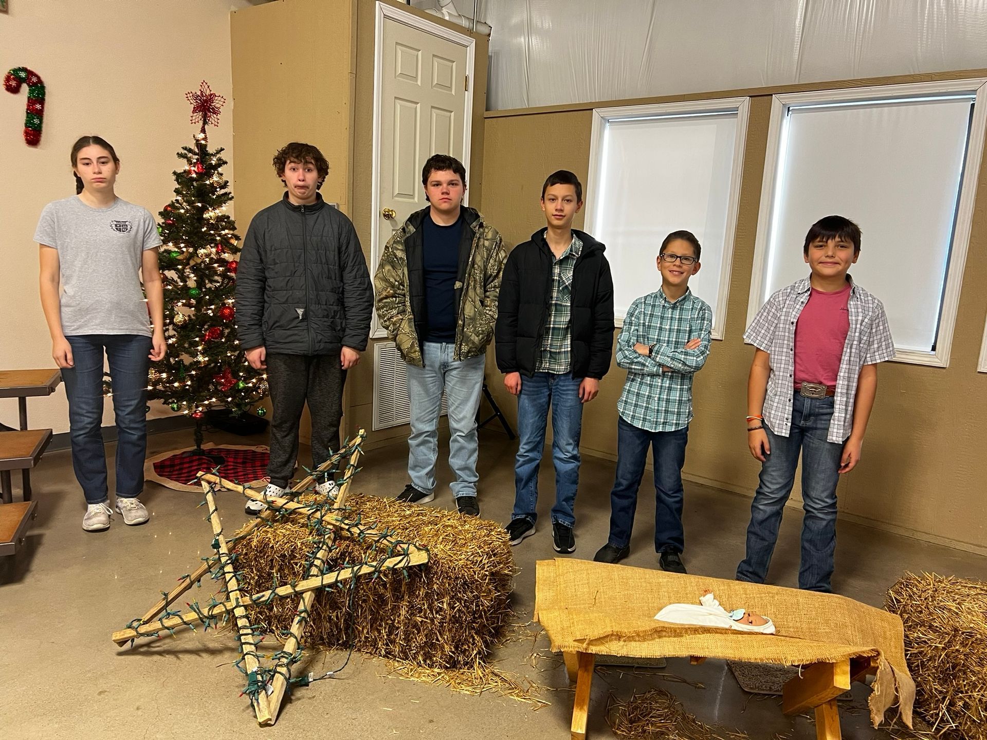 Six people stand in a room decorated for Christmas, near a star and hay bales.