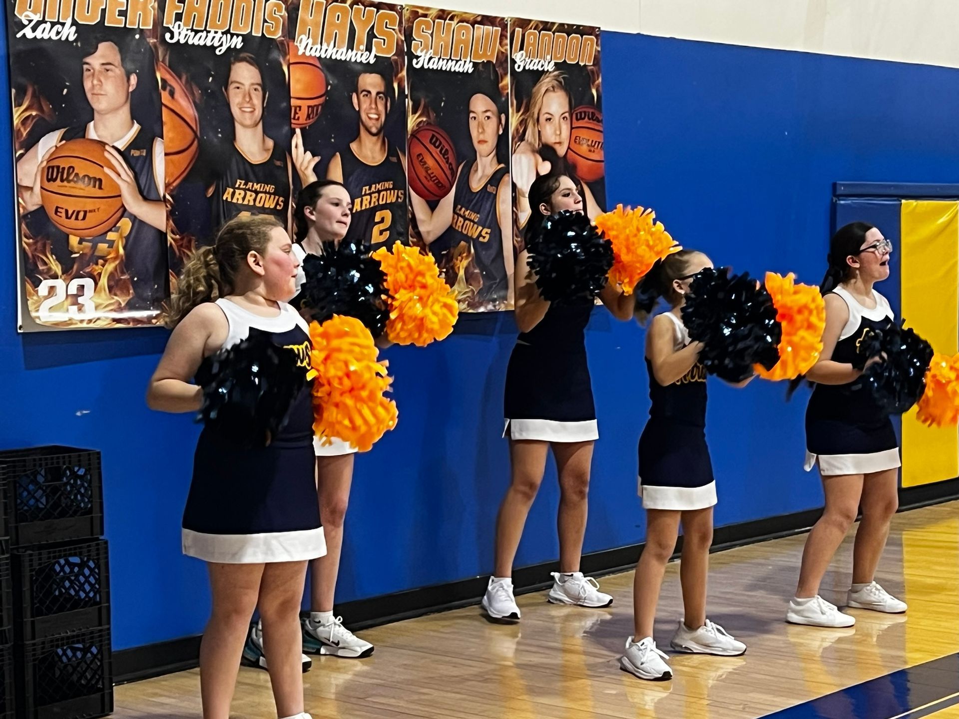 Cheerleaders in black and white uniforms perform with orange pom-poms in a gymnasium.