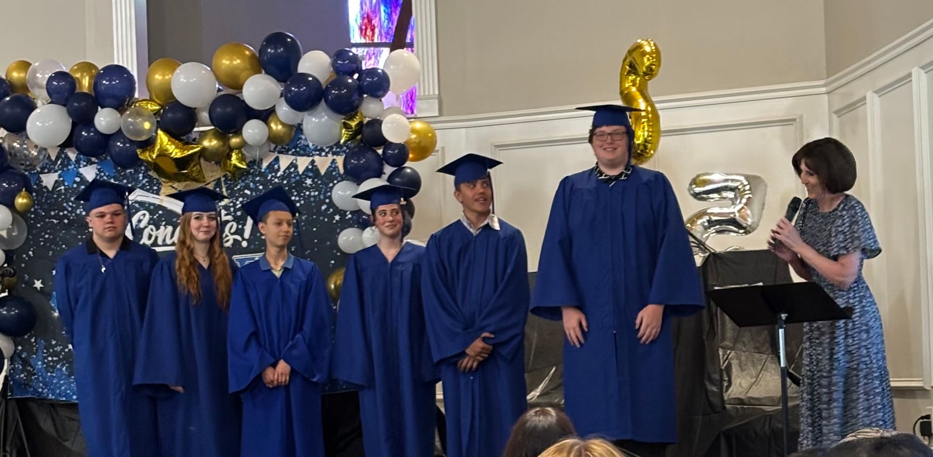 Graduates in blue gowns at ceremony; woman speaking into microphone. Balloons in background.