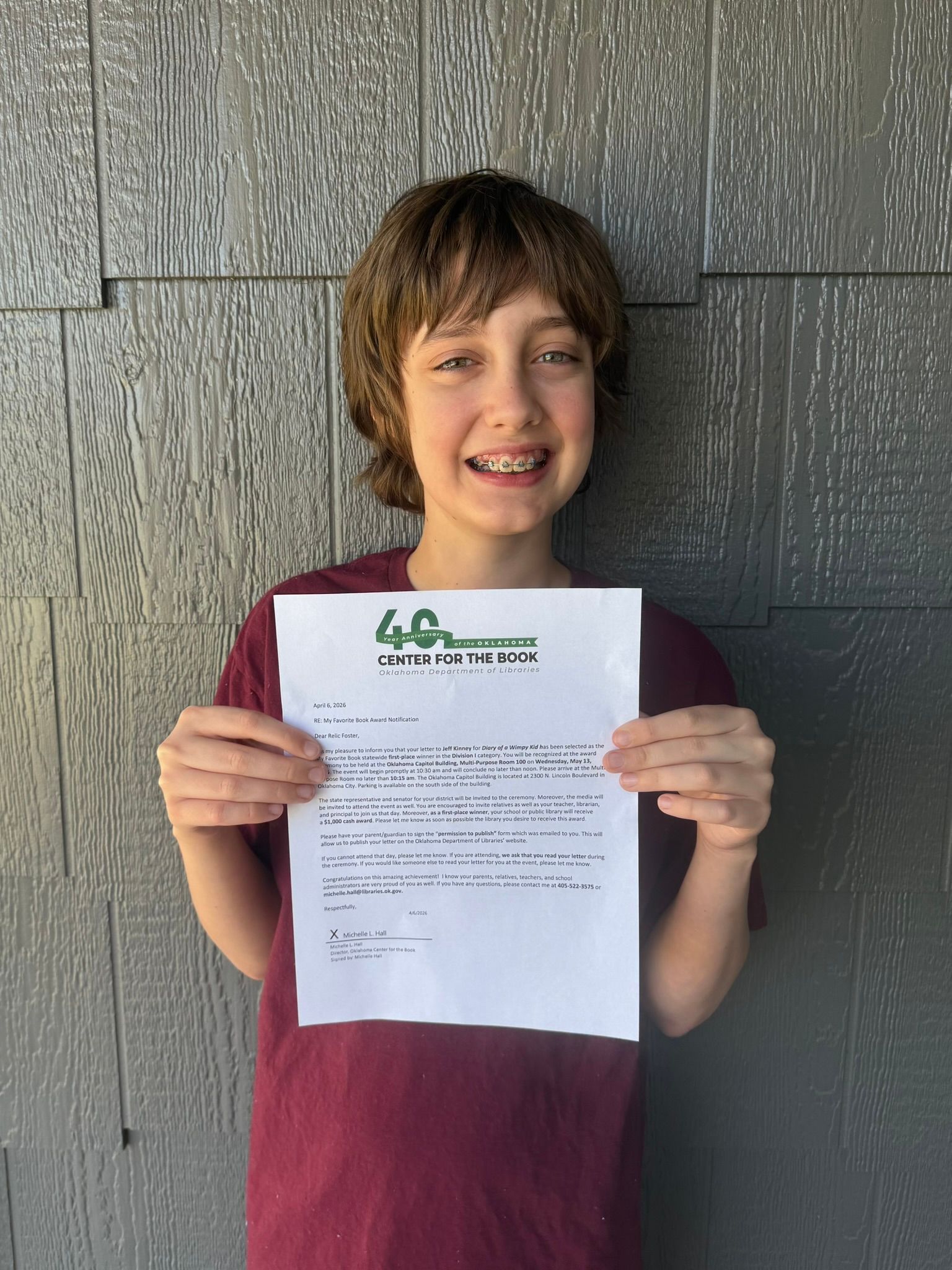 Person holding up a document in front of a gray wall, wearing a maroon shirt and braces.