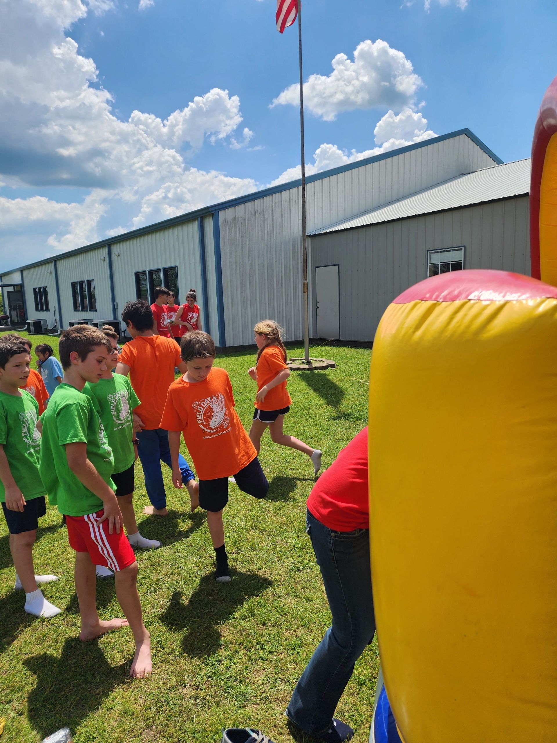 Children playing outside near a building with an American flag. Some are wearing orange and green shirts.