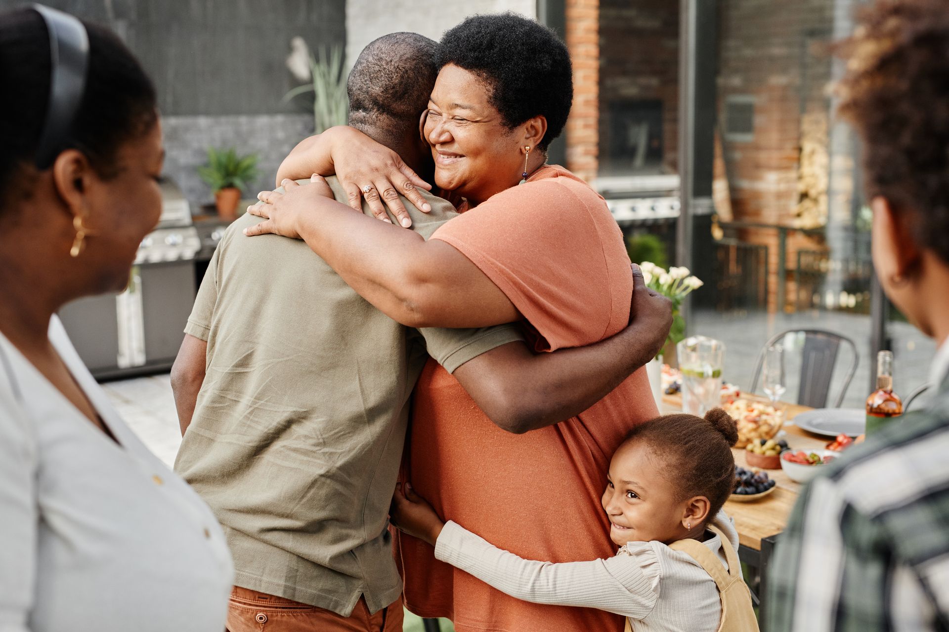 A group of people embrace in an outdoor setting, with a child hugging an adult’s waist while two adults share a hug.